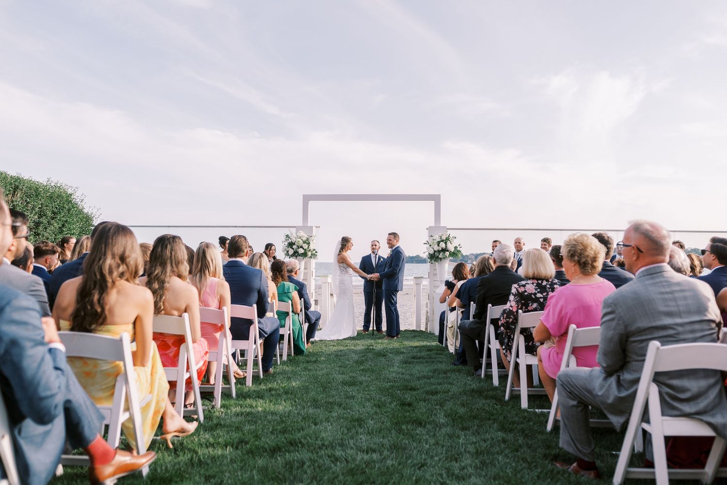 Wedding ceremony guests seated on white chairs on a lawn overlooking the water during an outdoor summer celebration.