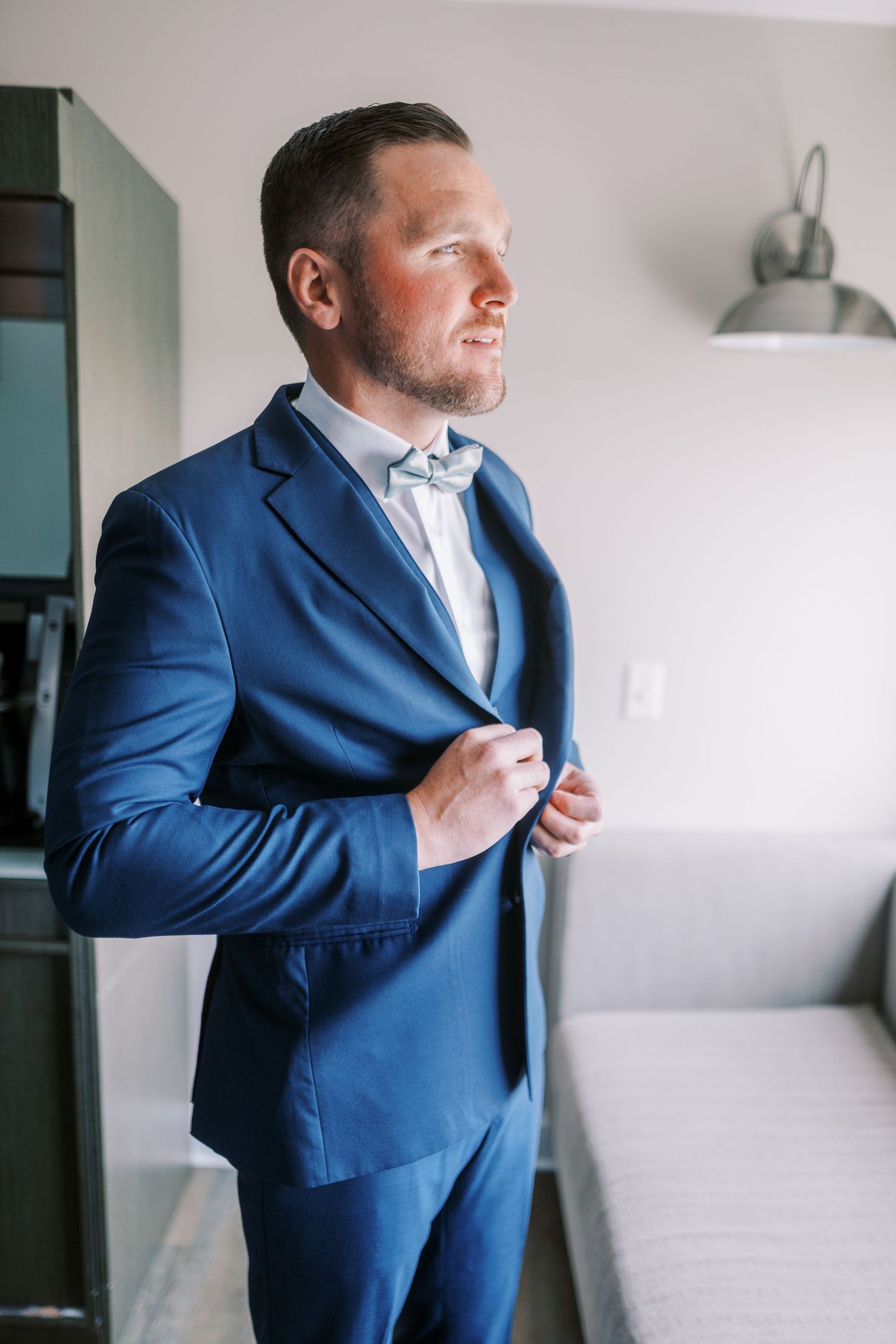 A person in a navy blue suit jacket adjusts their collar while getting dressed in a light-filled room.