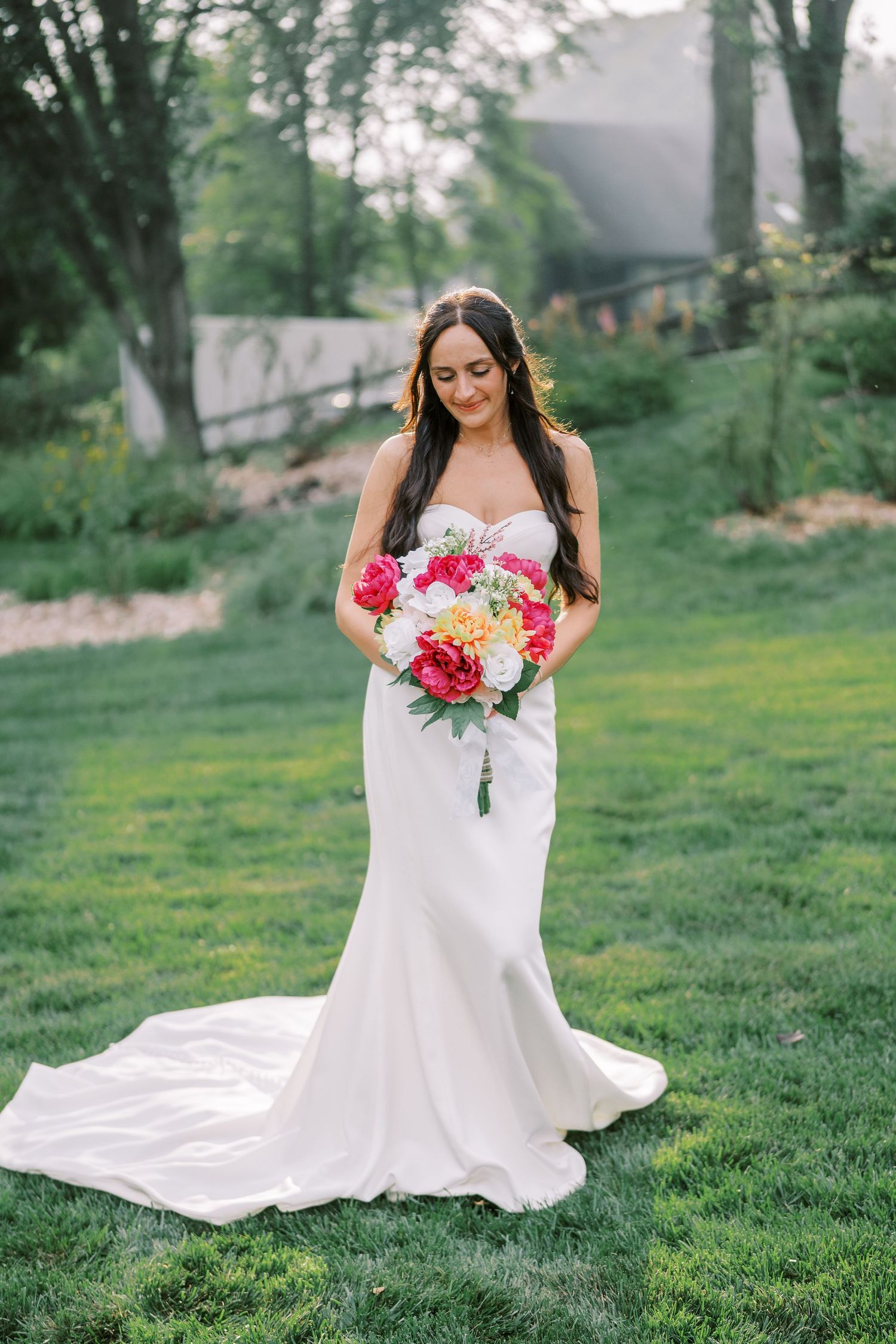 A bride in a white wedding gown sits gracefully on green grass while holding a vibrant pink and red bouquet.