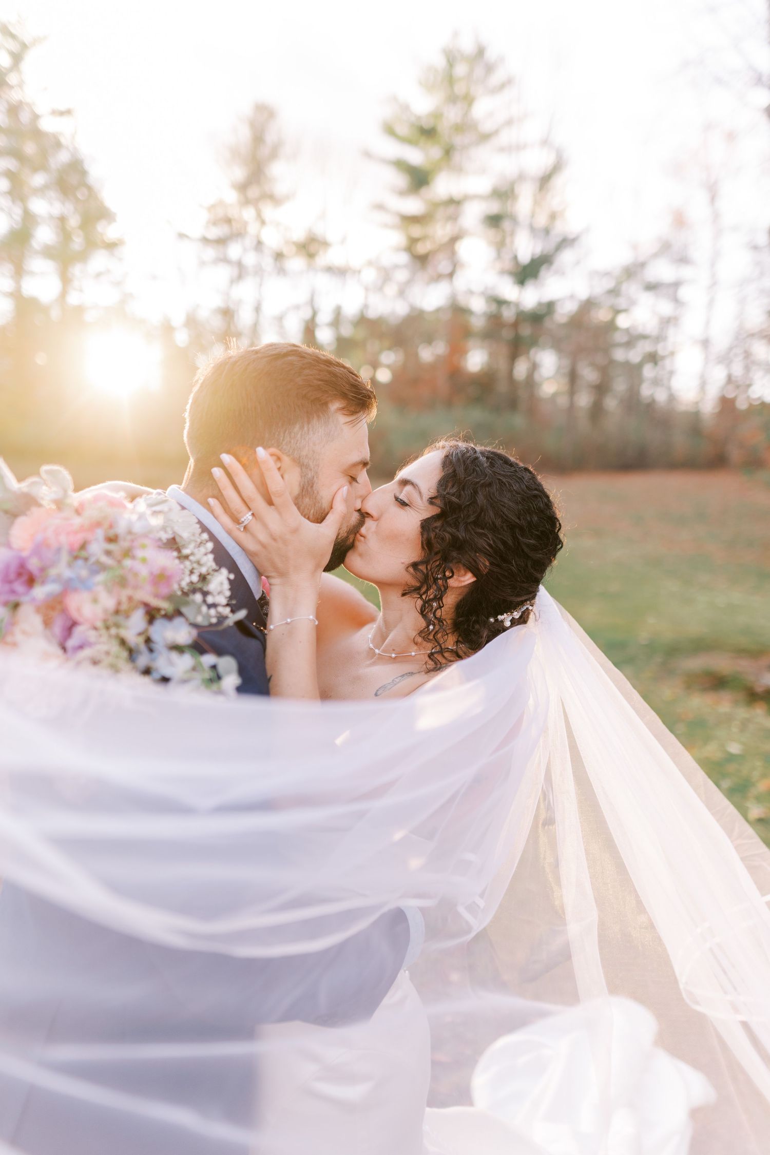 A romantic wedding couple shares a tender kiss while a flowing white veil catches the sunlight in an outdoor setting.