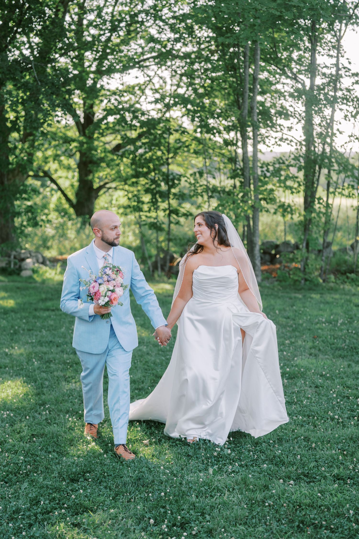 Couple walking on grass in wedding attire with light blue suit and white gown.