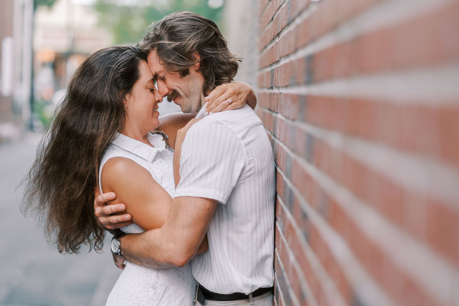 A romantic couple shares an intimate embrace against a brick wall in an urban setting.