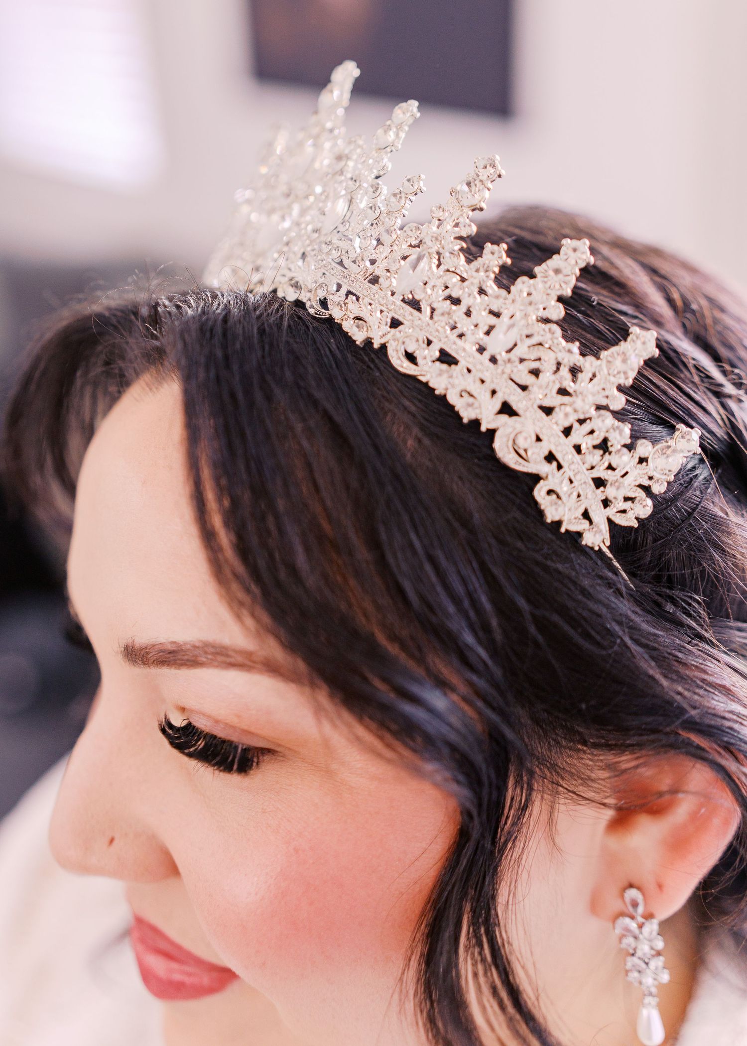 Delicate pearl and crystal bridal tiara crown adorns dark hair with matching earrings and soft pink makeup.