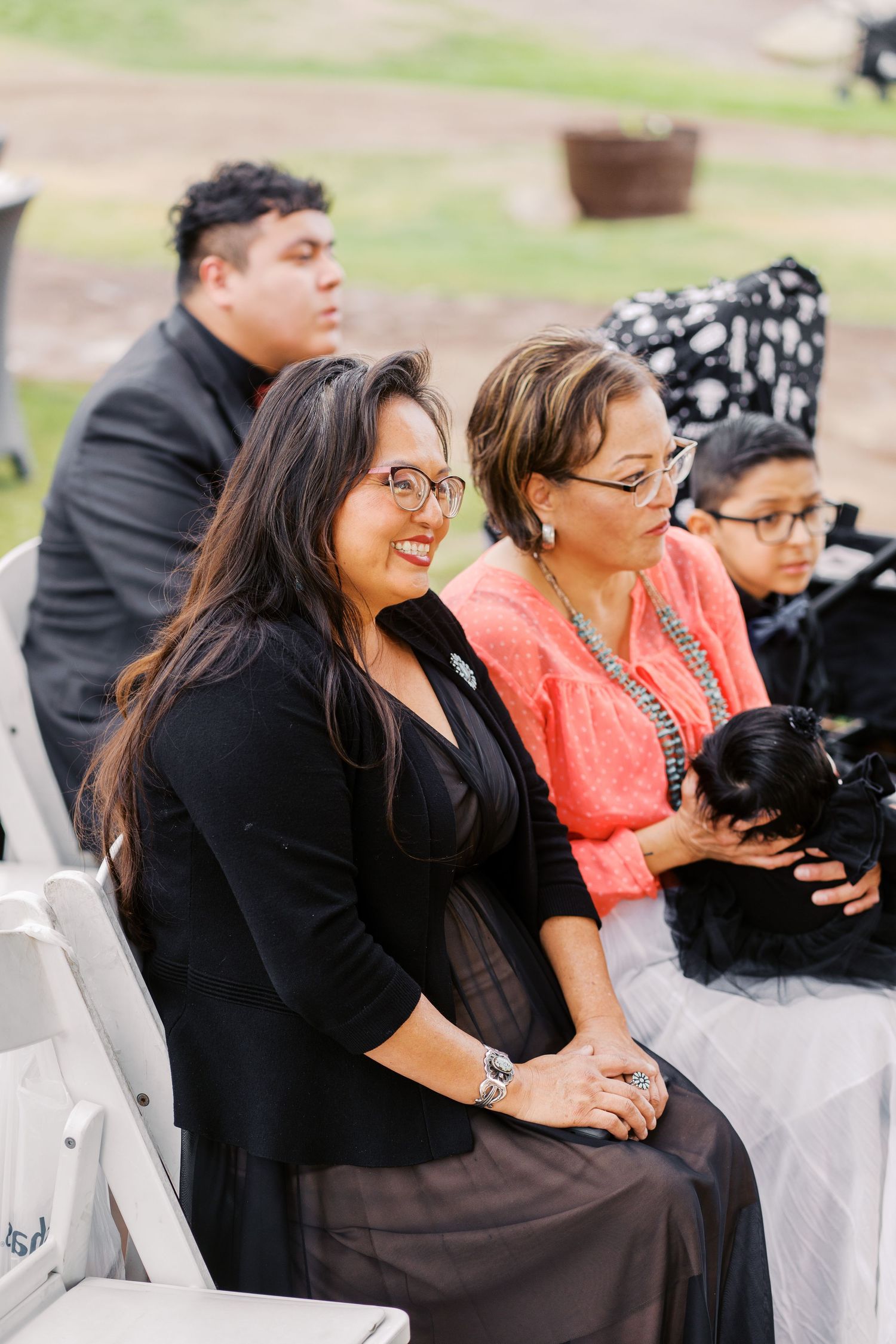People sitting in folding chairs at an outdoor event, wearing formal attire including a black blazer and coral dress.
