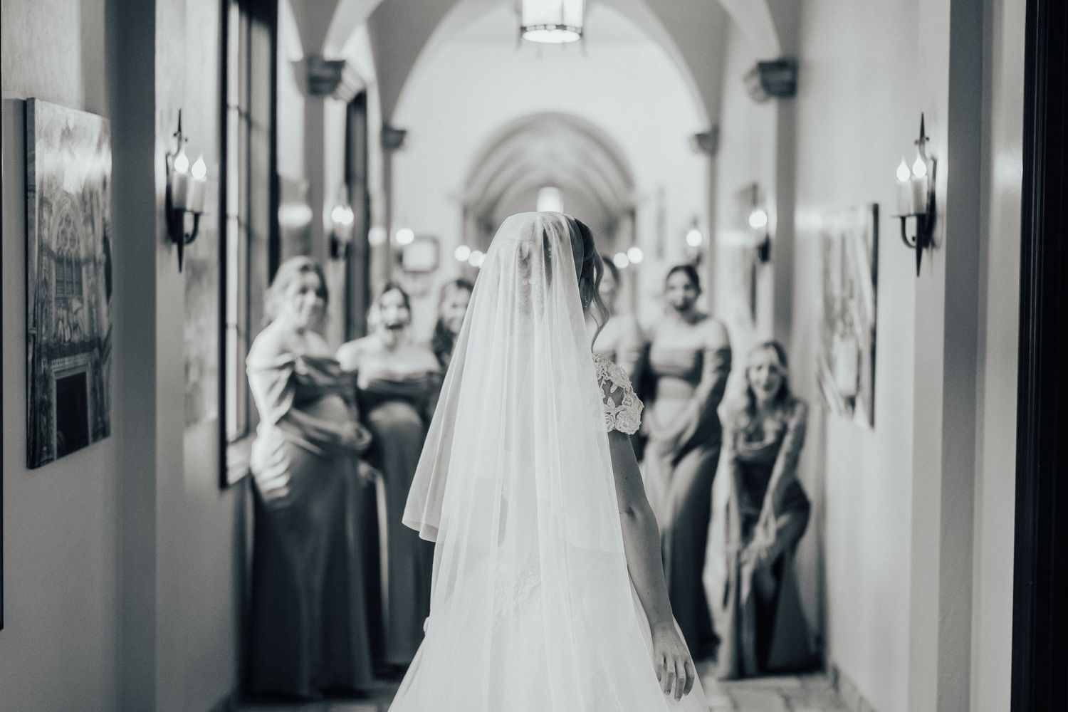 A bride in a flowing white veil stands in an elegant arched hallway while bridesmaids look on from behind.