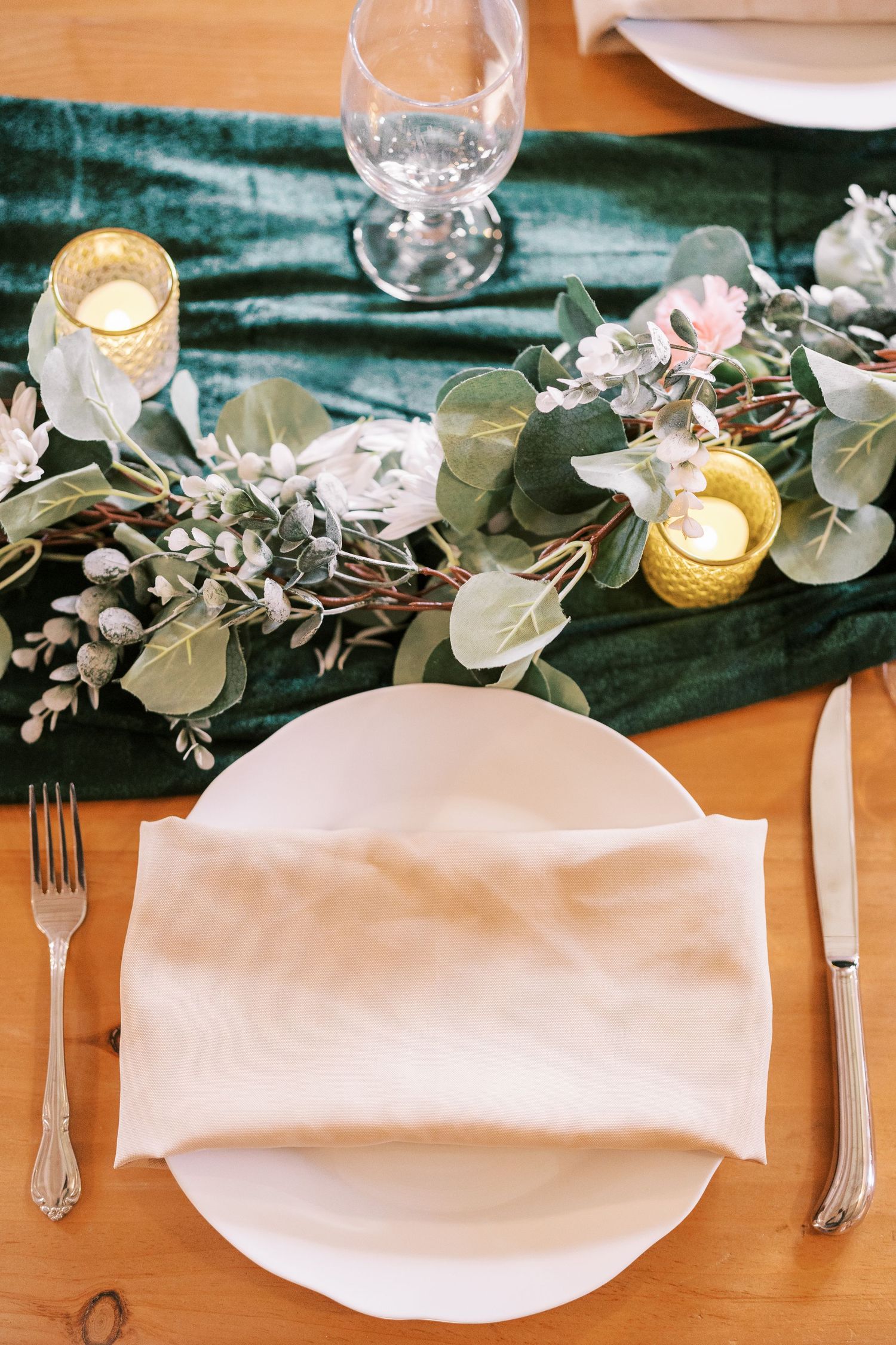 Elegant table setting with white plate, eucalyptus garland, candlelight and emerald green velvet runner on wooden surface.