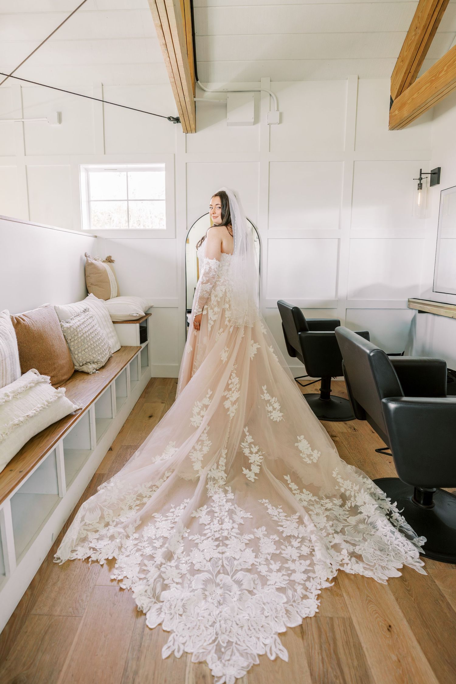 Elegant white wedding dress with intricate lace train displayed in a bright, minimalist room with wooden floors.