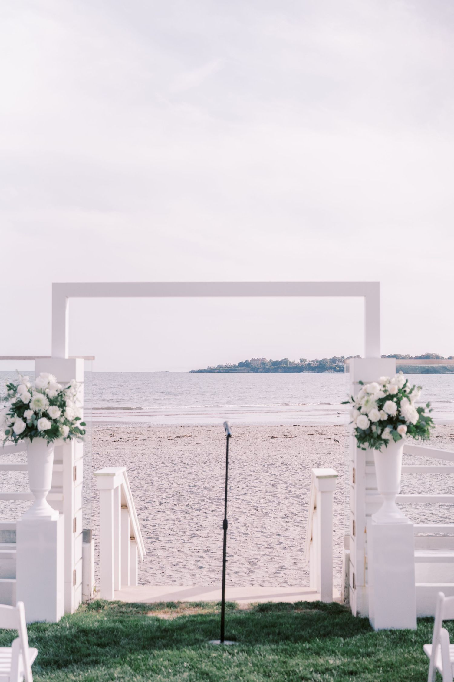 White wedding arch decorated with floral arrangements stands on a beach with white chairs and ocean view in the background.
