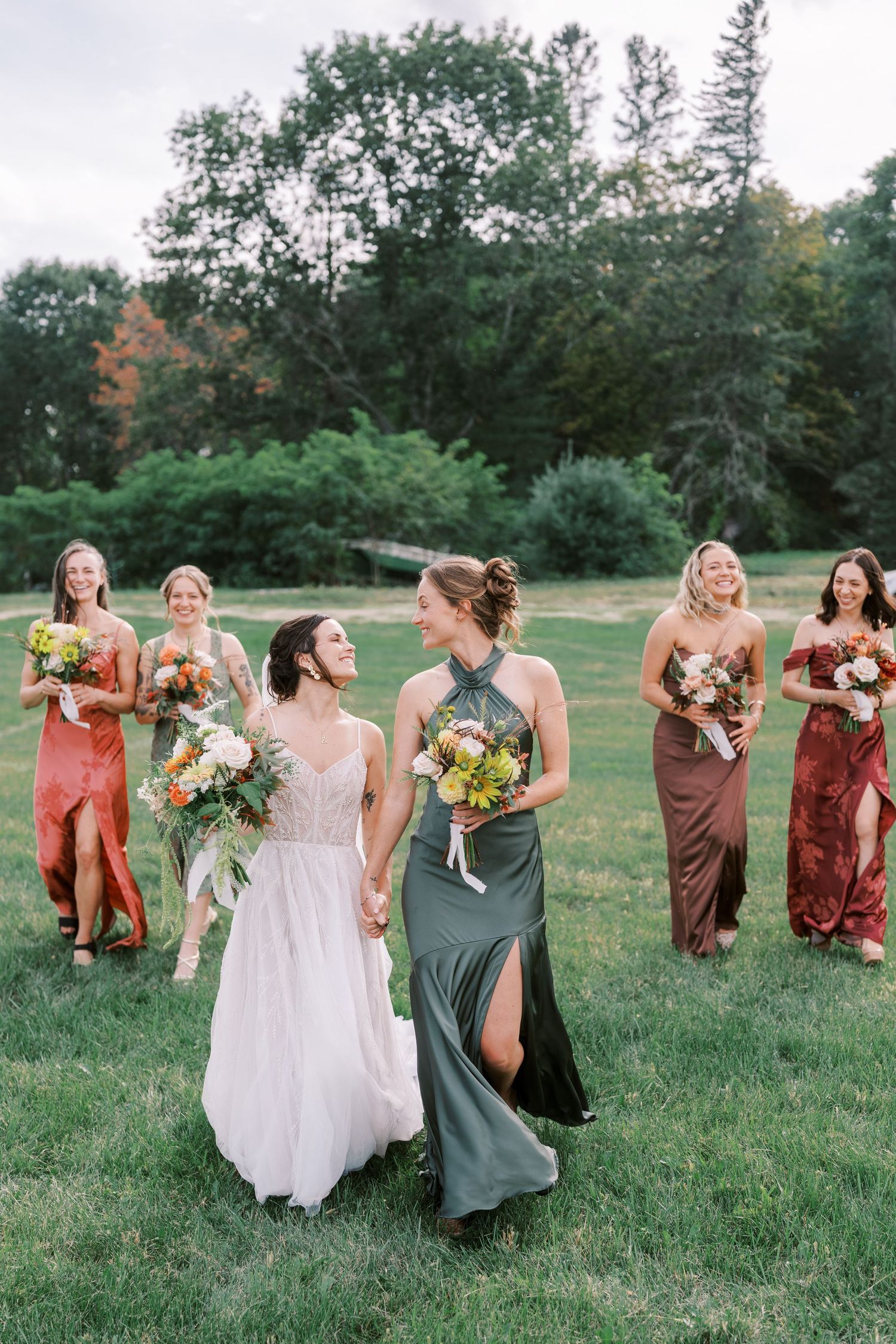 Bridesmaids in elegant dresses carrying bouquets walk across green lawn.