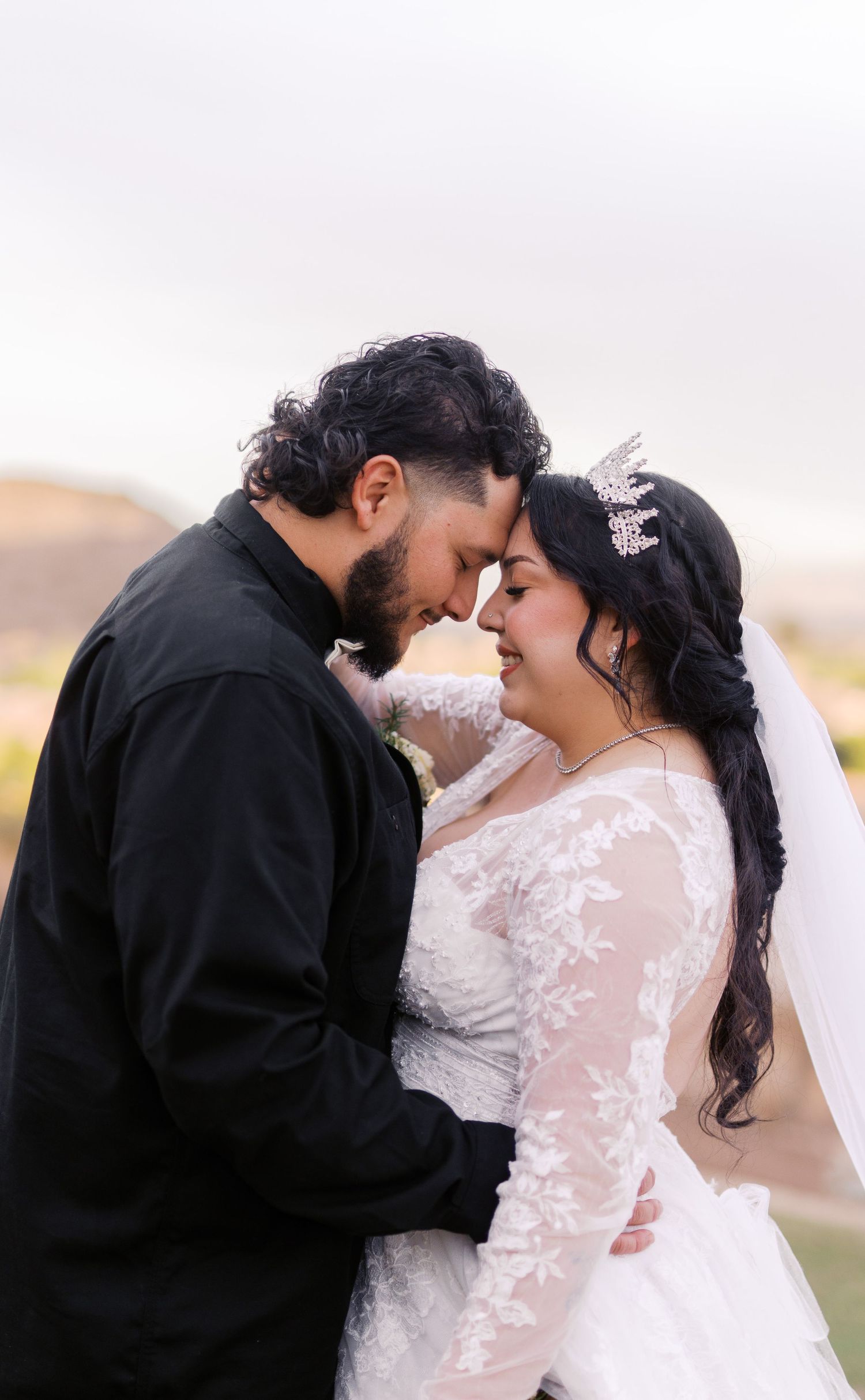 A tender moment between newlyweds as they share an intimate embrace outdoors, the bride wearing a lace gown and delicate headpiece.