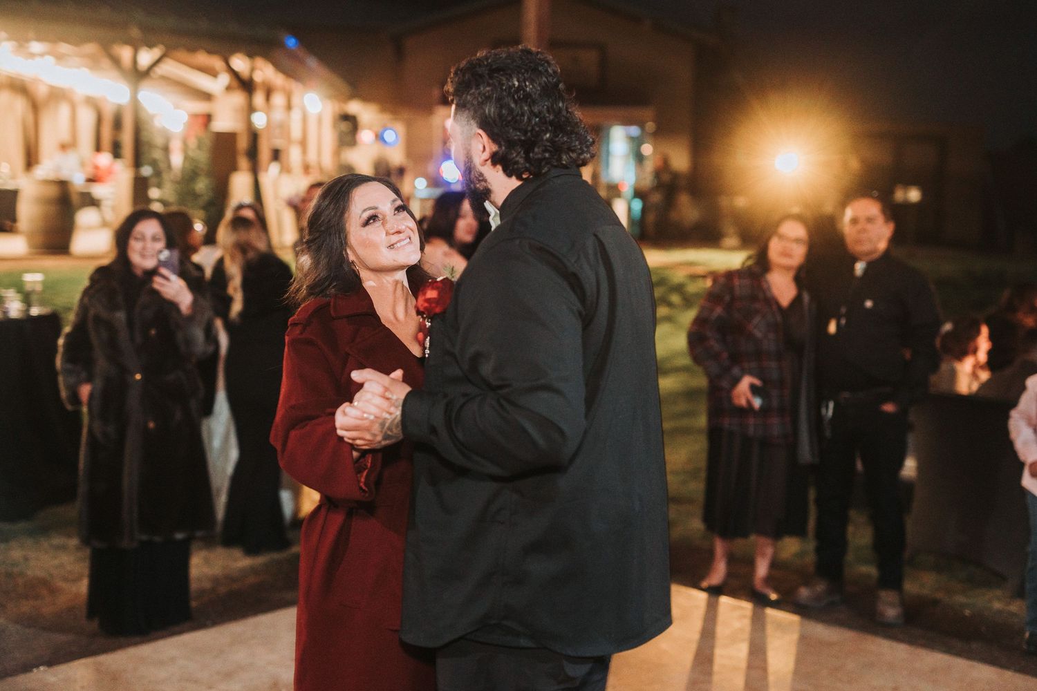 Two people in winter coats share a romantic moment outside at night while others watch in the background.