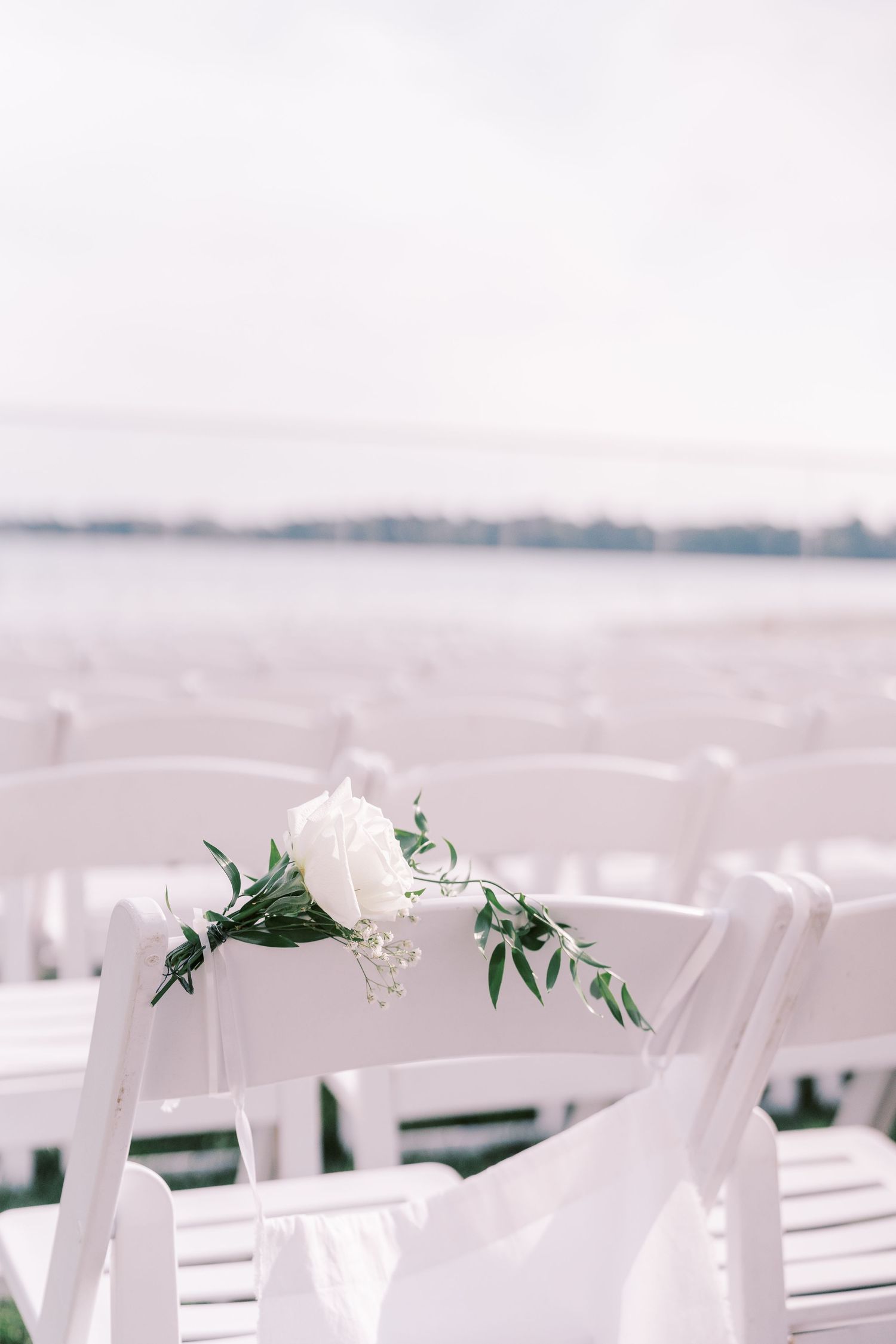 White folding chairs decorated with delicate greenery and flowers set up for an outdoor wedding ceremony by the water.