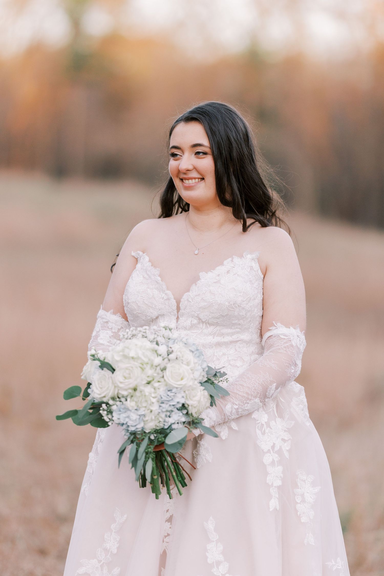 A bride in a white lace dress holds a delicate bouquet of white flowers and greenery in an outdoor setting.