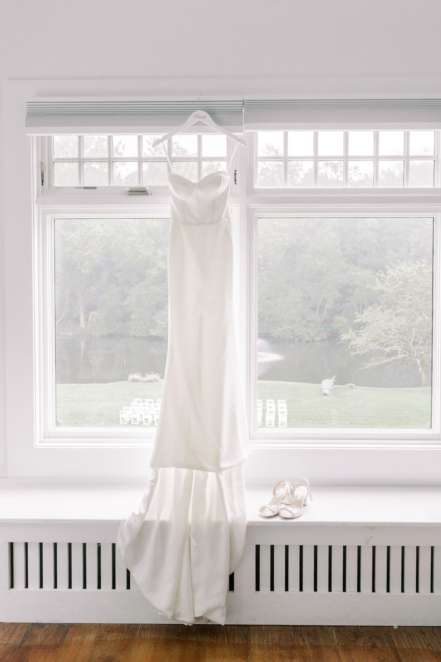 An elegant white wedding dress hangs by a bright window with white heels placed beneath it.