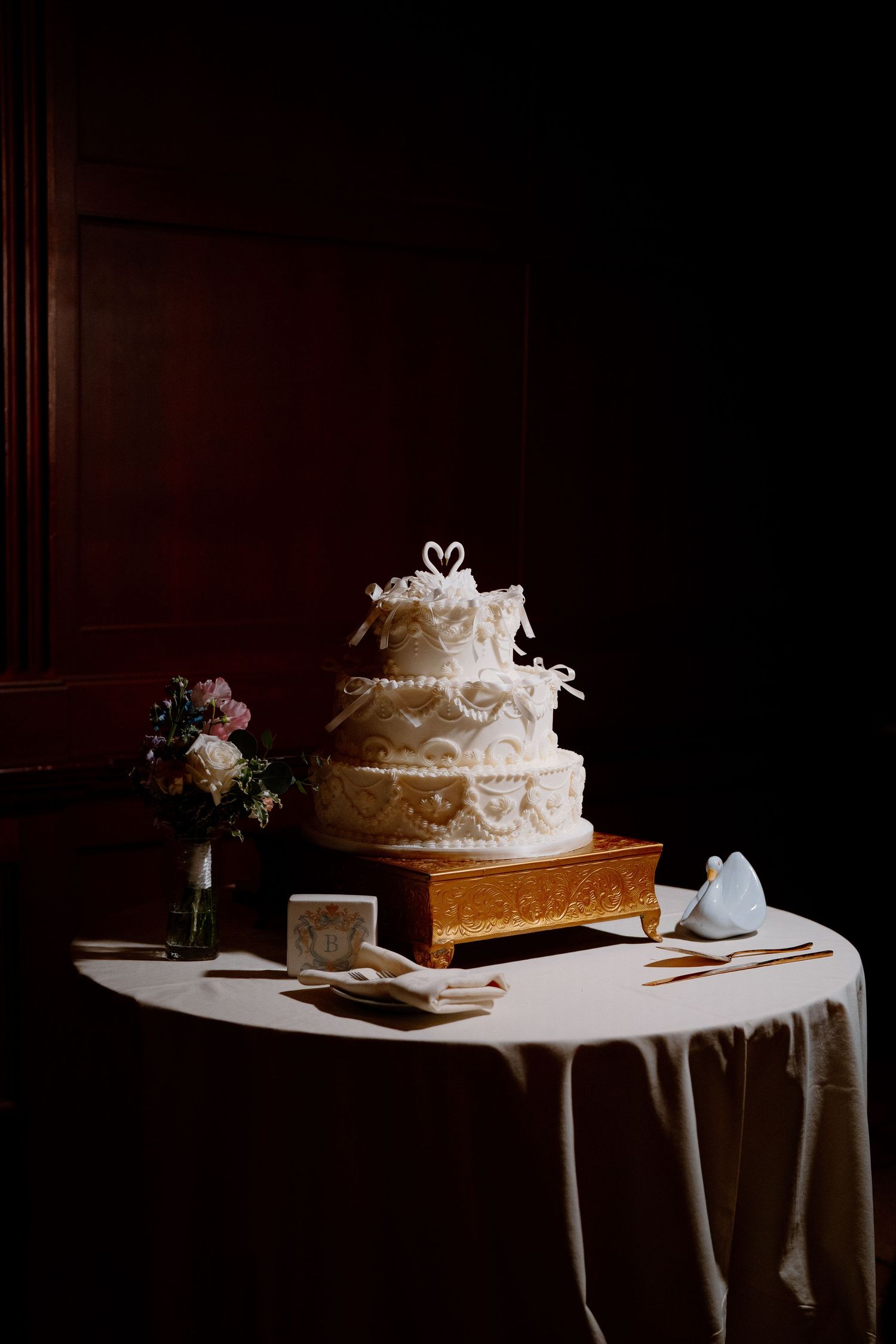 Elegant white wedding cake with swan topper displayed on white tablecloth in dramatic lighting.