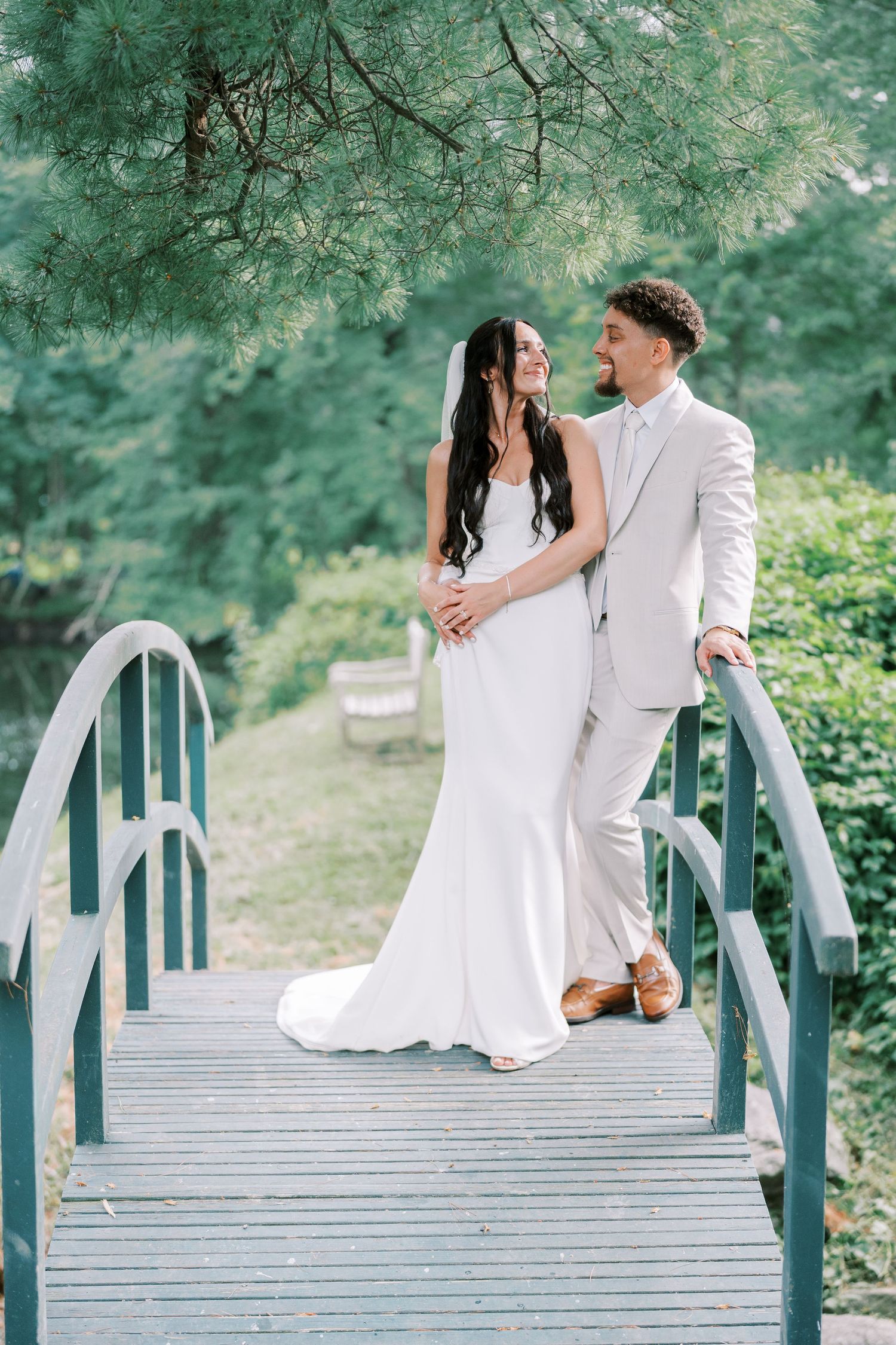 A couple shares an intimate moment on a wooden bridge surrounded by lush green trees.
