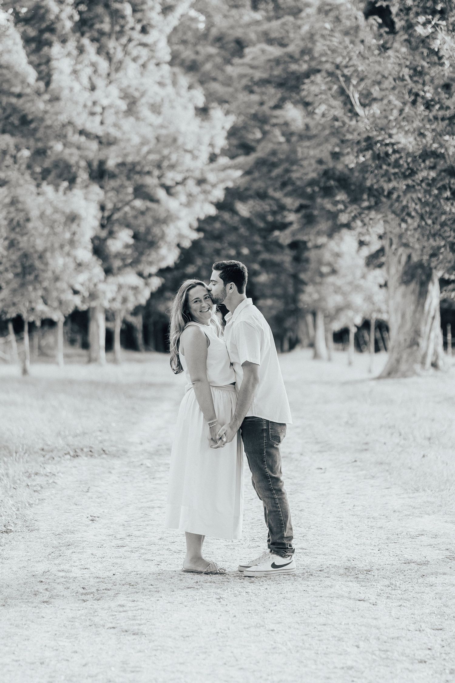 Black and white portrait of couple standing close together on a dirt path surrounded by trees.