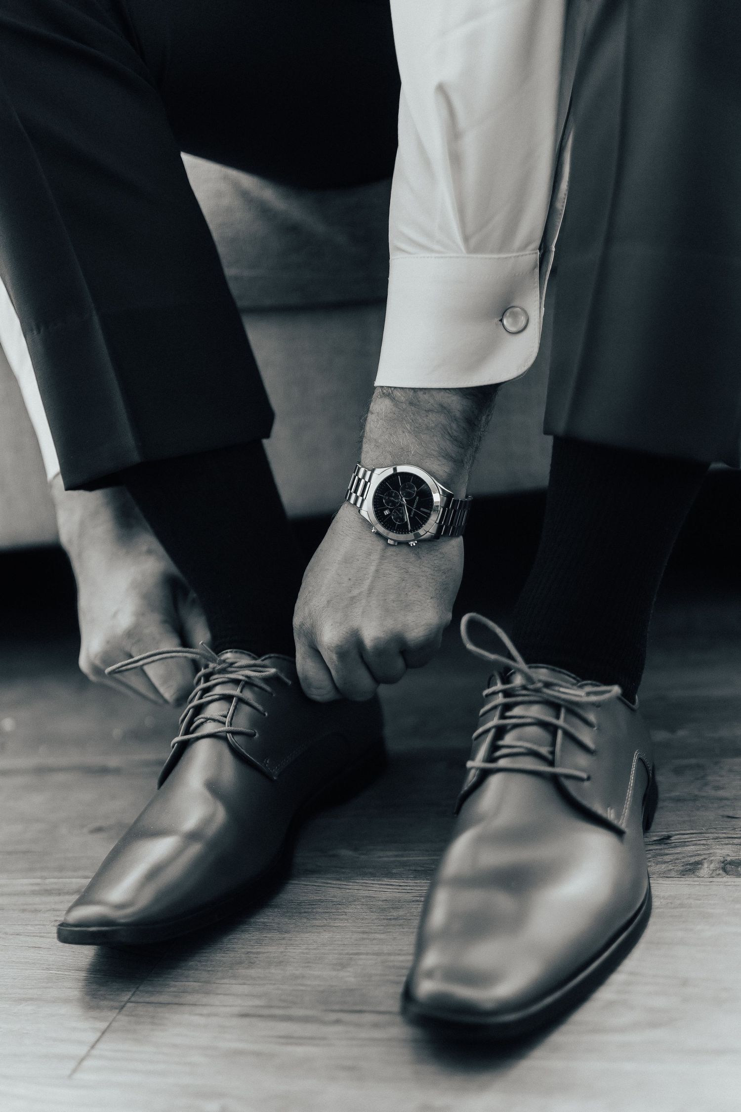 Close-up view of black leather dress shoes and wrist watch on person wearing white shirt and dark suit pants.