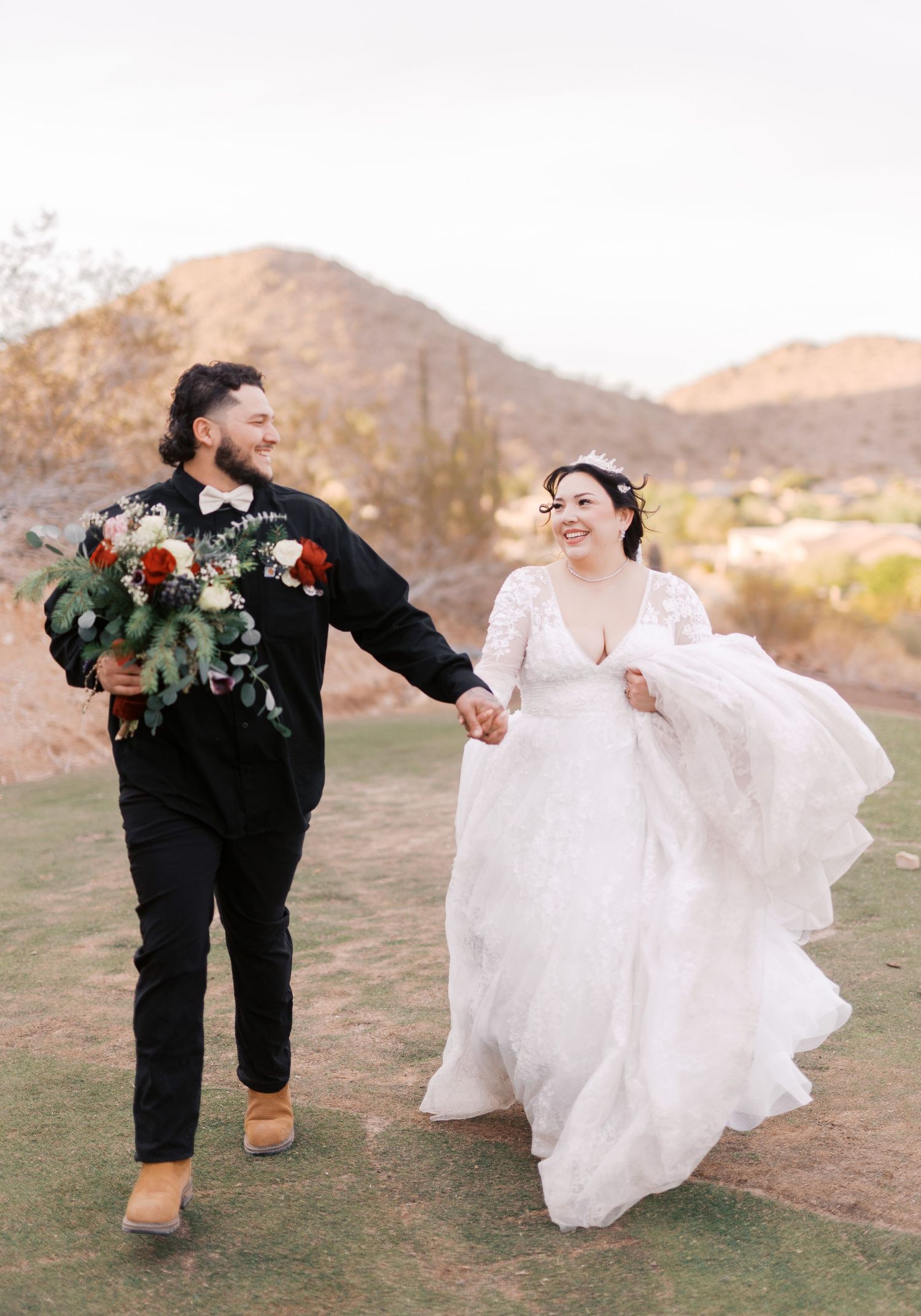 Joyful newlyweds walk hand in hand through a desert landscape with mountains, the bride's white dress flowing in the breeze.