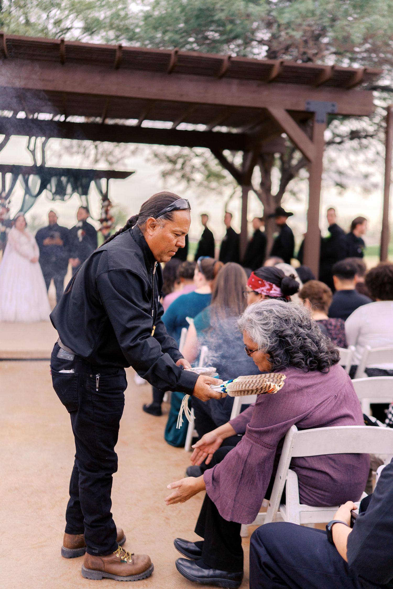 A person wearing dark clothing interacts with an elderly visitor seated at an outdoor covered gathering space.