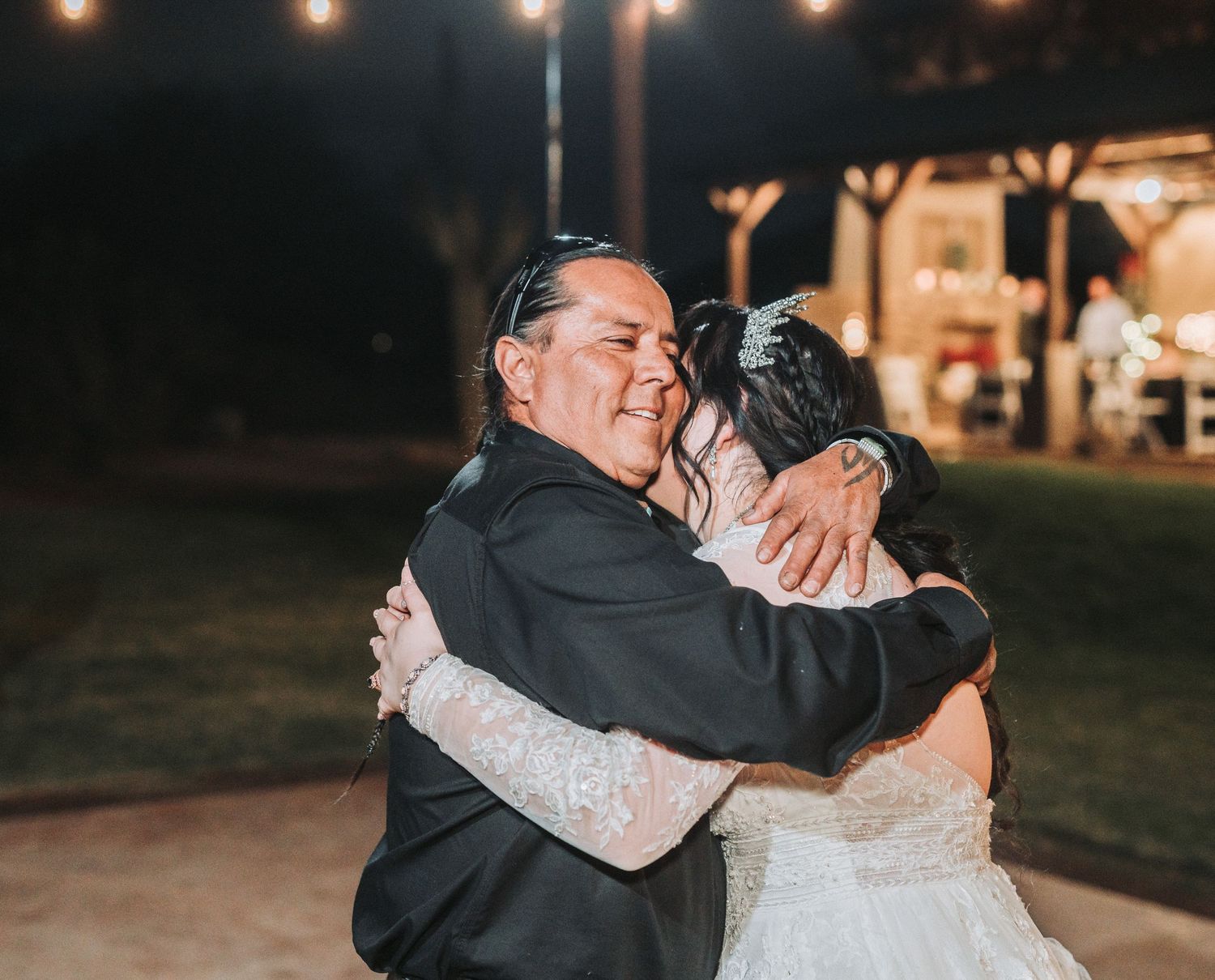 A couple shares an emotional embrace during an evening outdoor wedding reception with twinkling lights in the background.