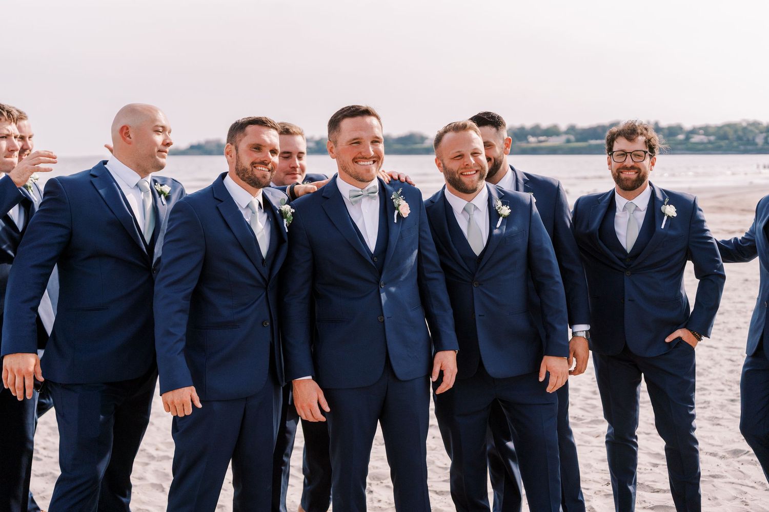 Groomsmen in matching navy suits laugh together while walking on a beach.