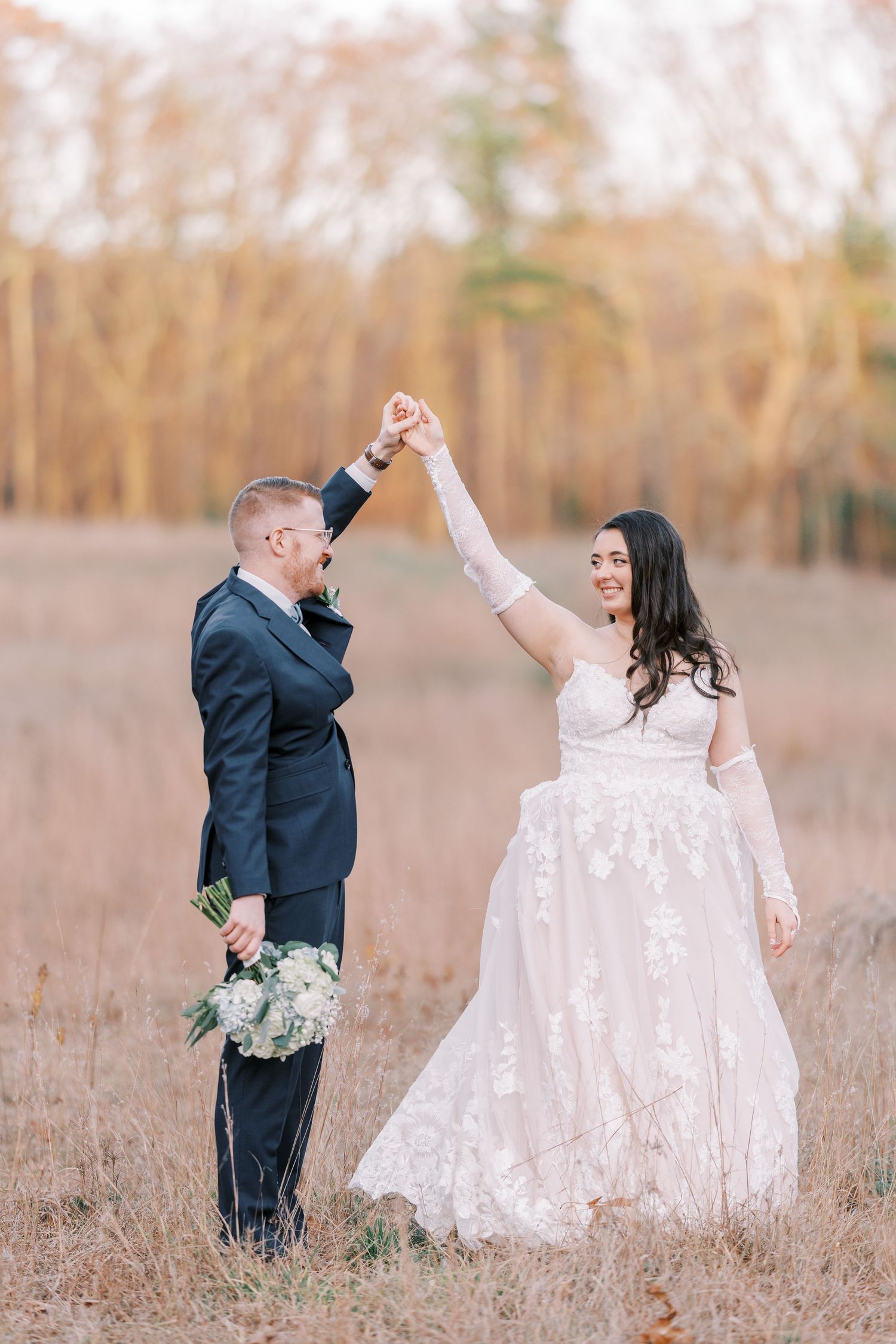 A couple celebrates their wedding day in a golden field with the bride in lace and groom holding a floral bouquet.