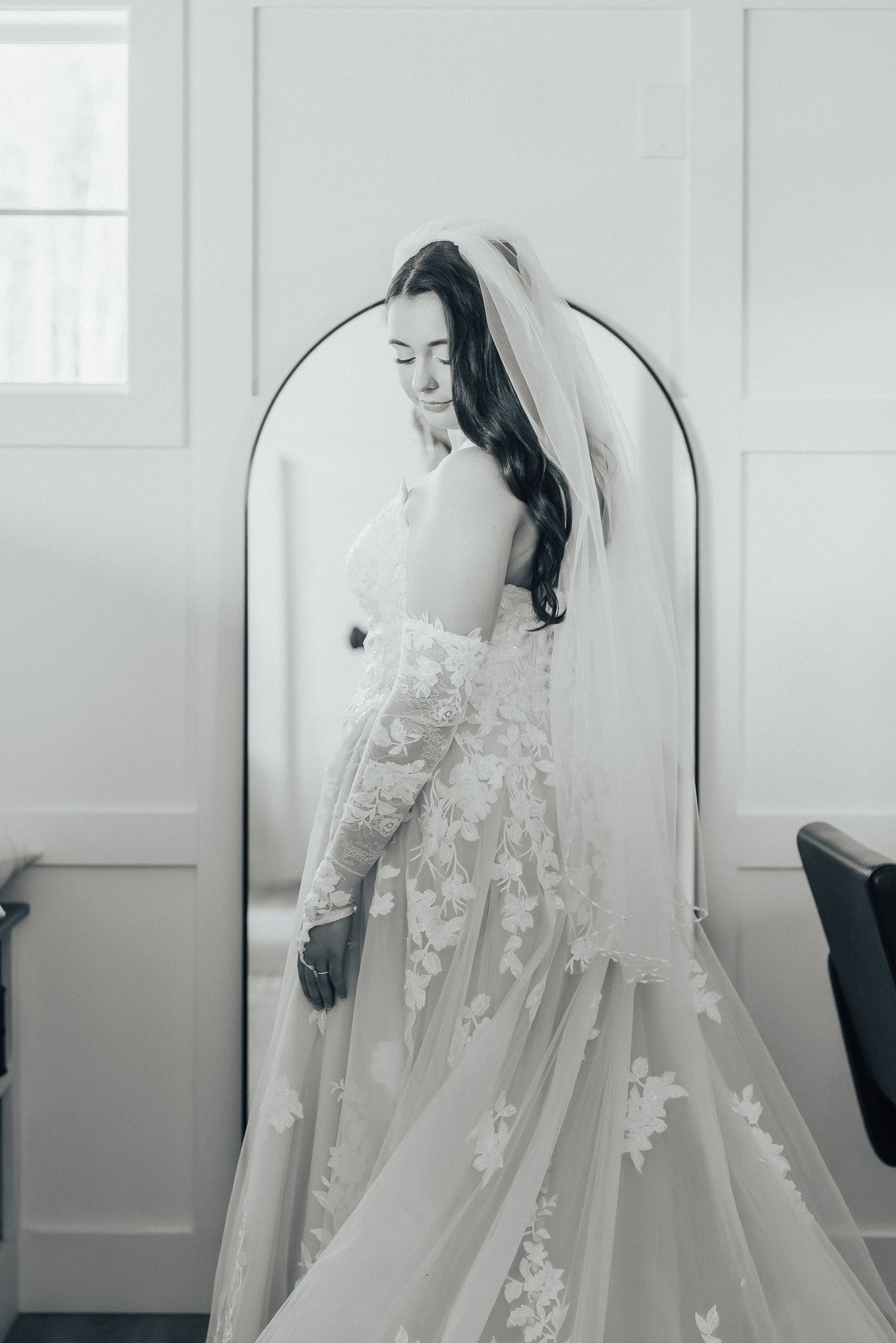 Elegant white wedding gown with delicate floral lace details and flowing veil photographed in an arched window frame.