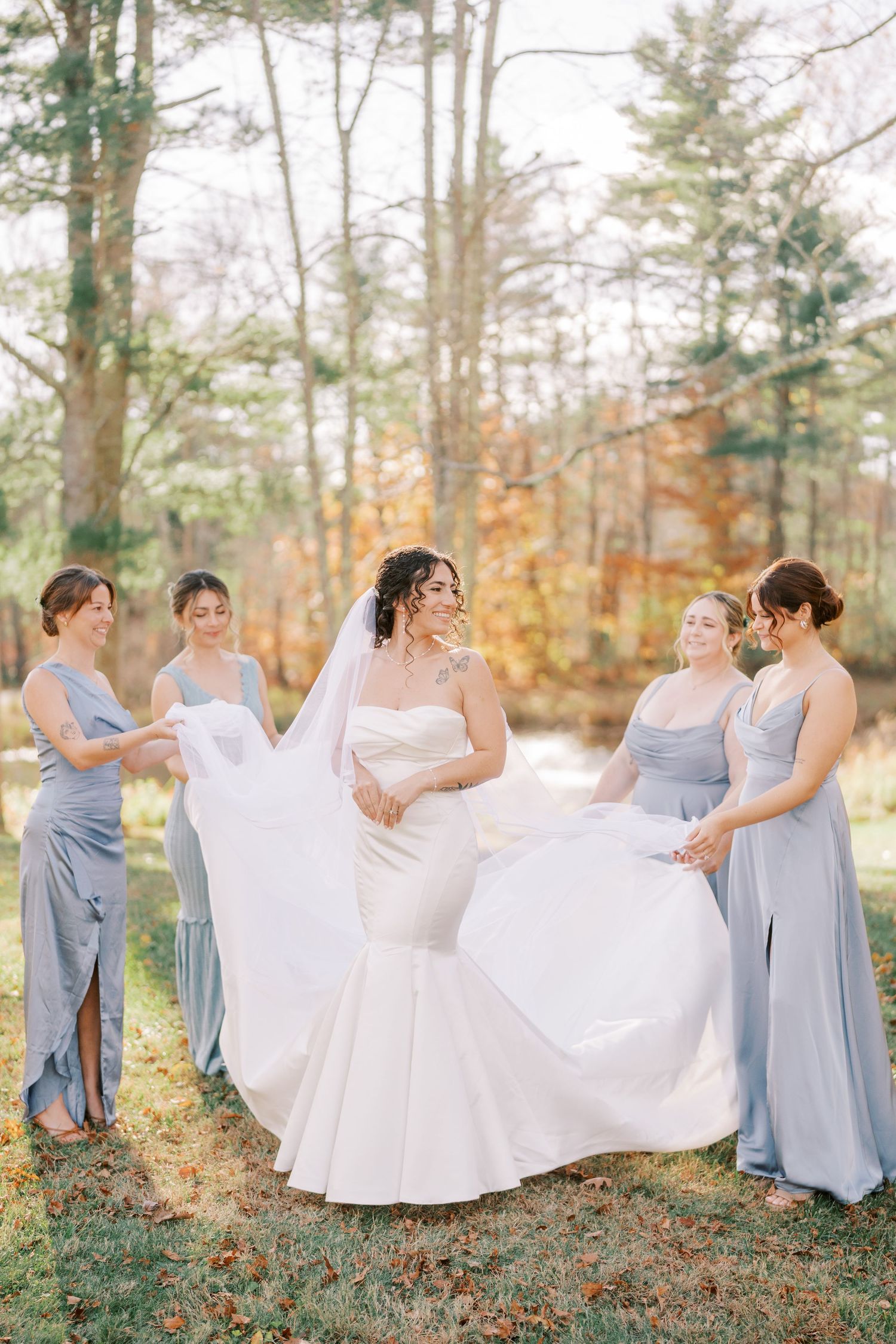 Bridesmaids in light blue dresses help bride with white wedding gown in autumn forest setting.