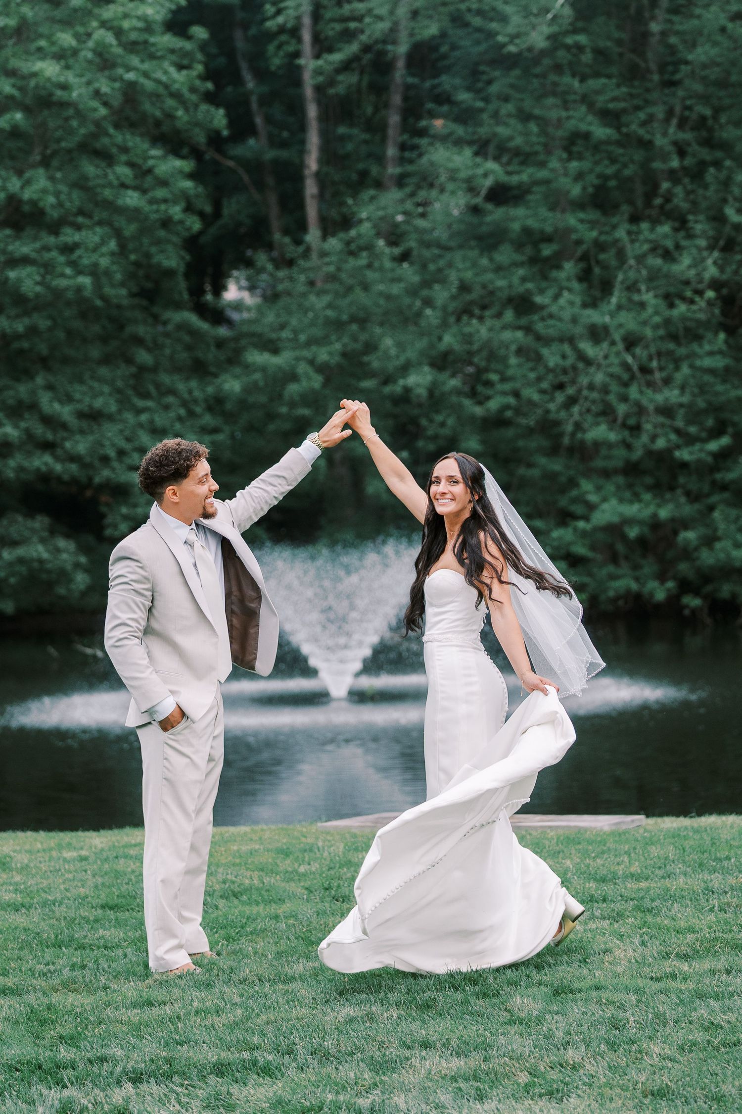 A joyful couple dances on manicured lawn with fountain and forest backdrop.