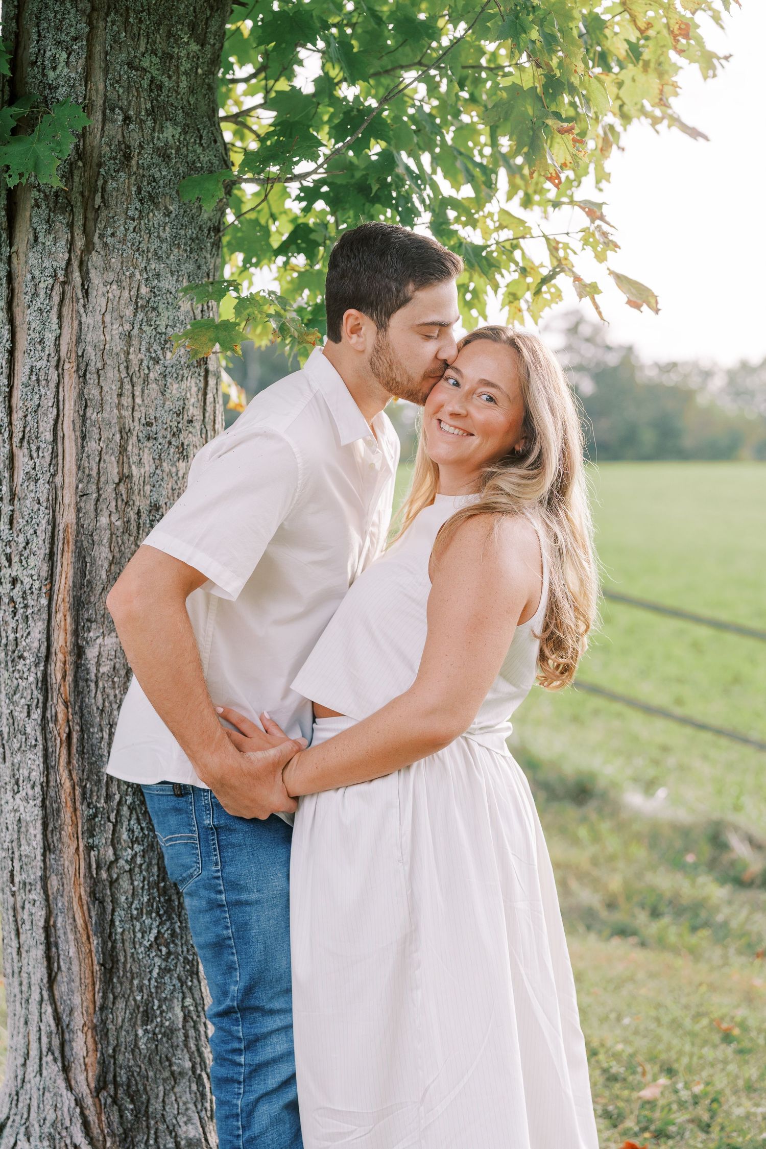 A couple in coordinated white outfits sharing a romantic moment against a large tree trunk.