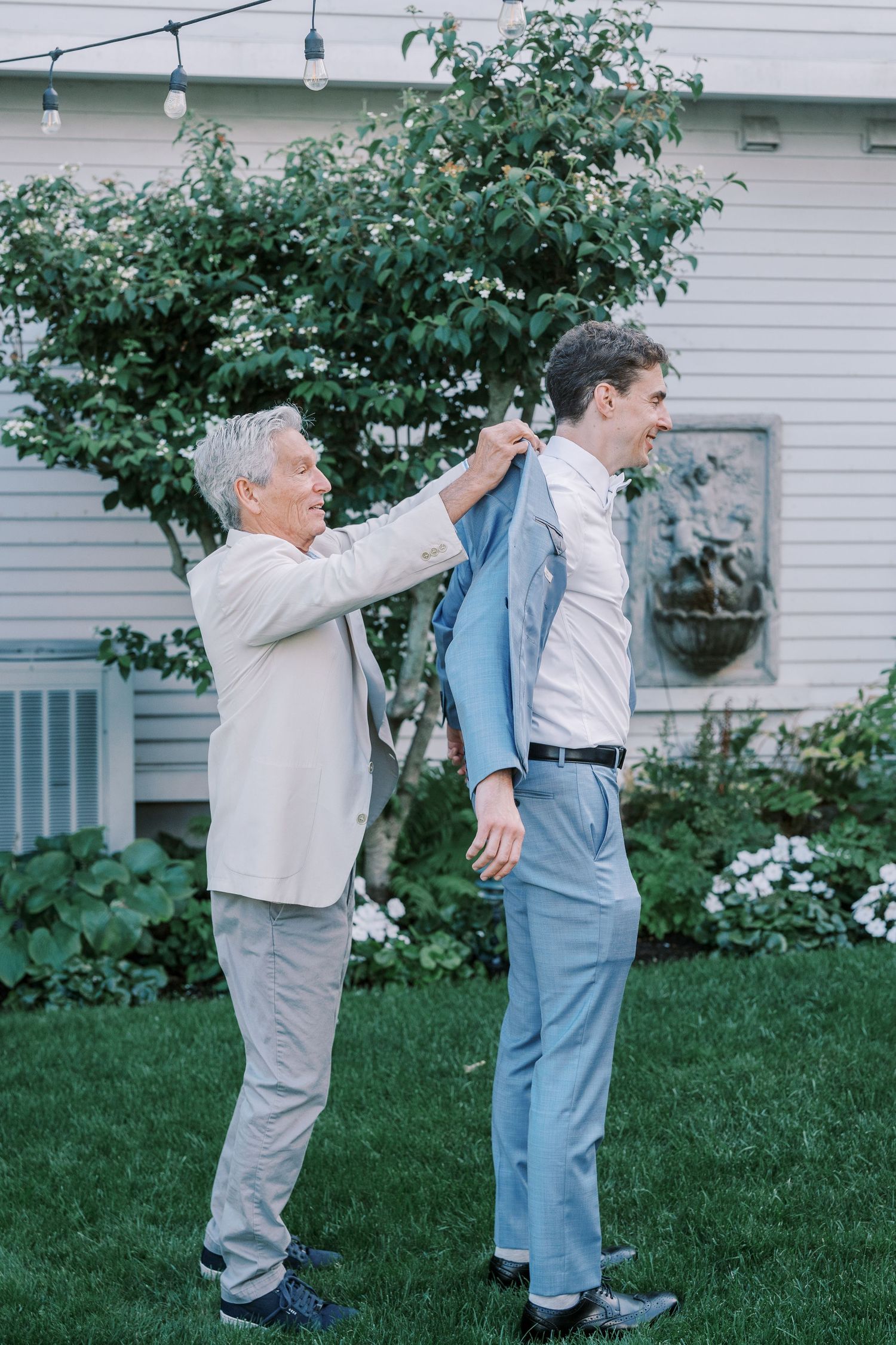 Two people in suits playfully interacting on a lawn in front of a white house.