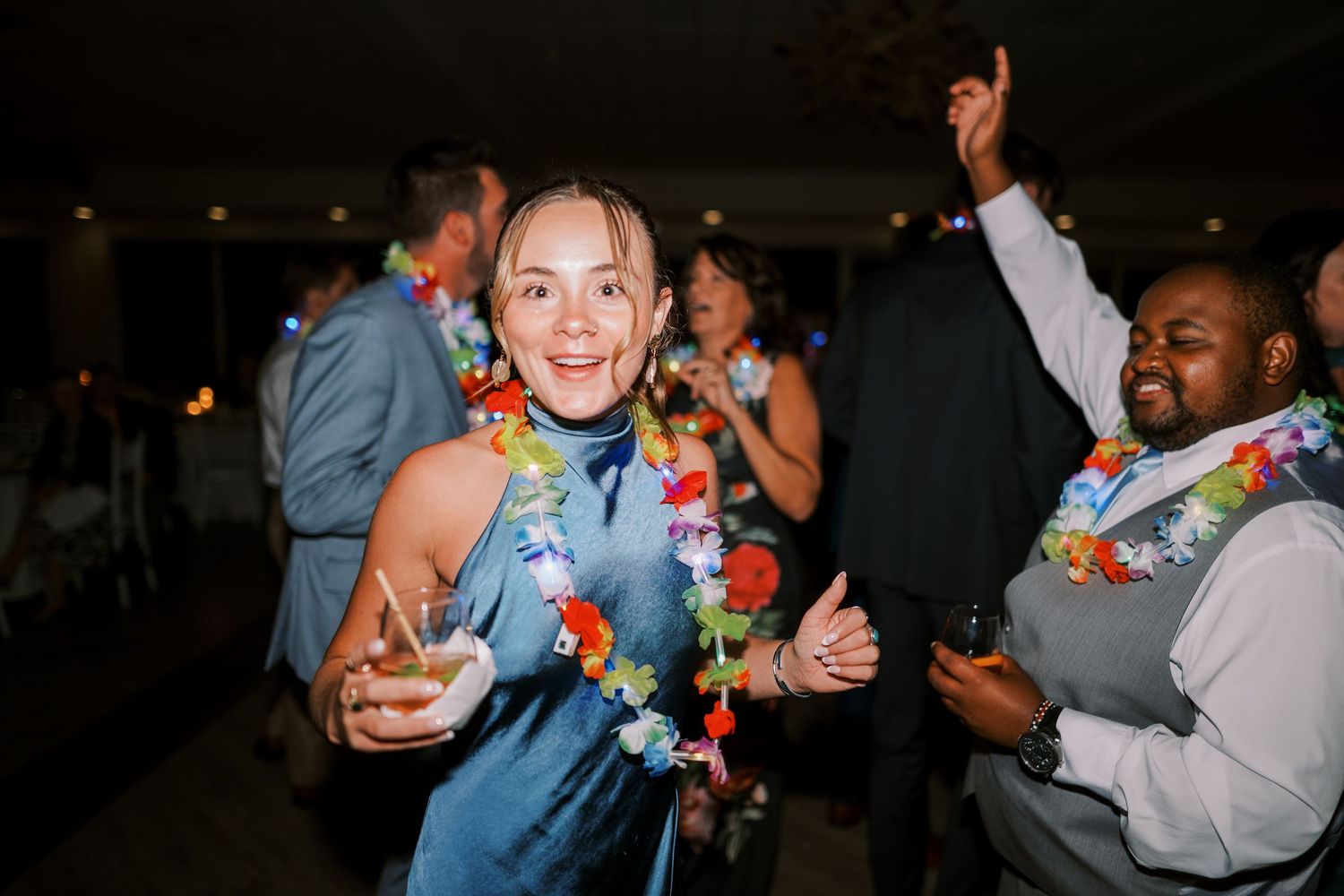 People wearing colorful Hawaiian leis and casual attire celebrate at a dimly lit indoor party gathering.