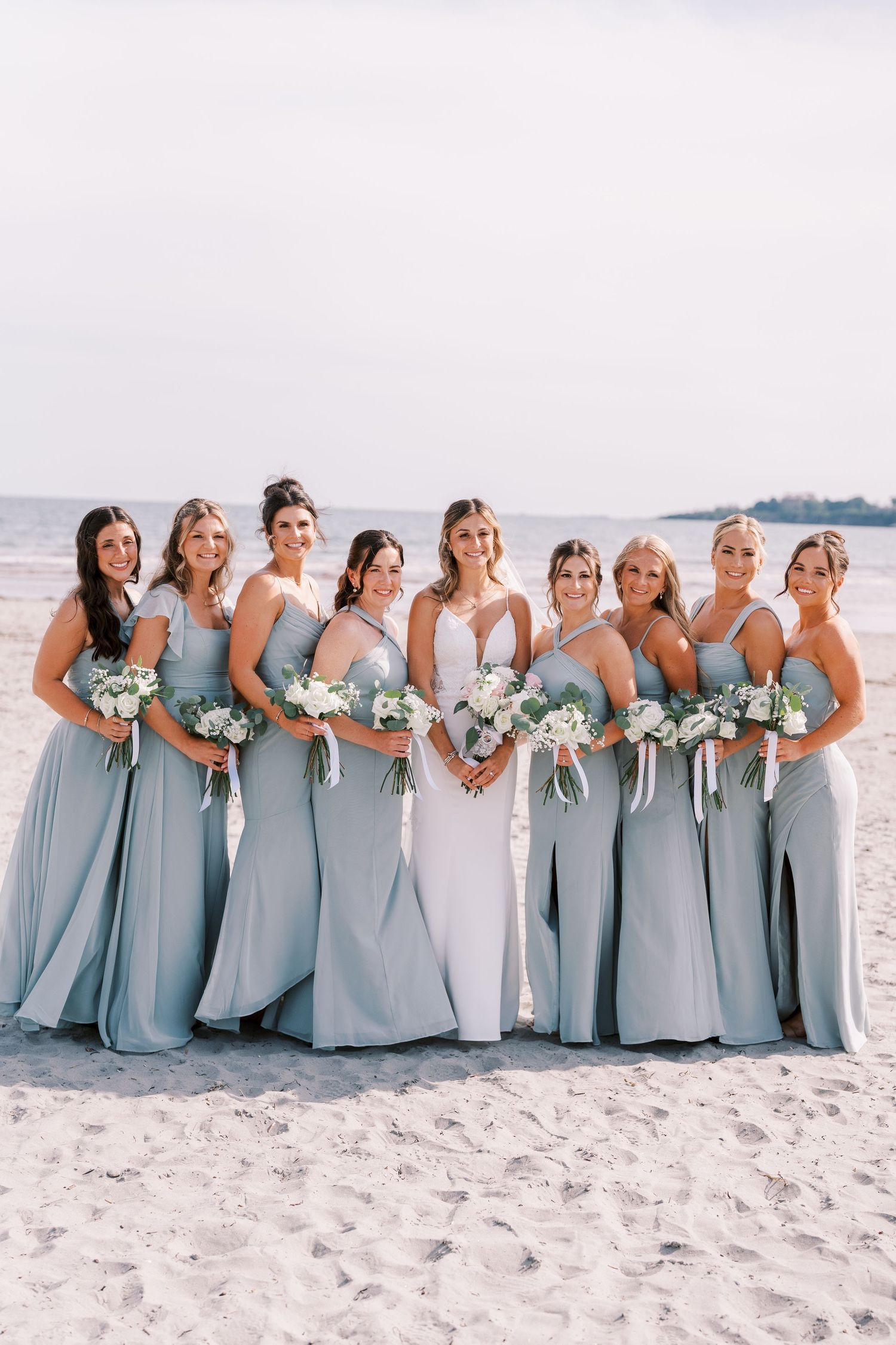 Bridesmaids in light blue dresses pose together on a sunny beach with white sand and ocean backdrop.