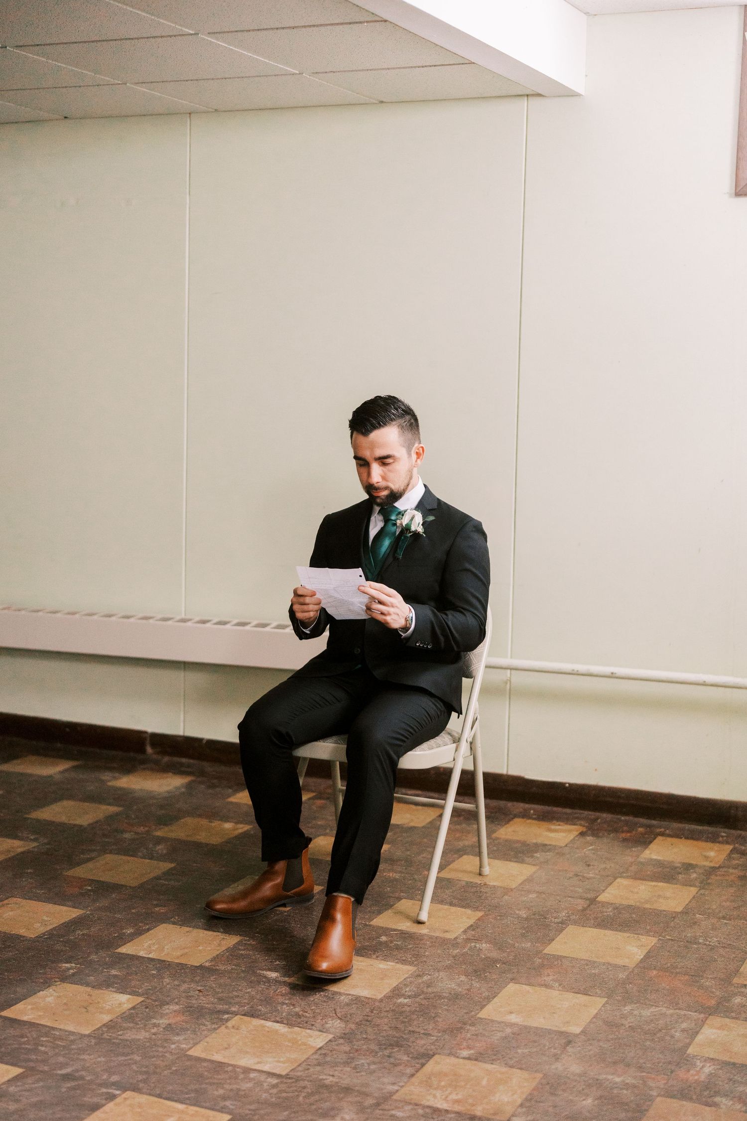 Groom in black suit and brown boots reads letter while seated on folding chair.