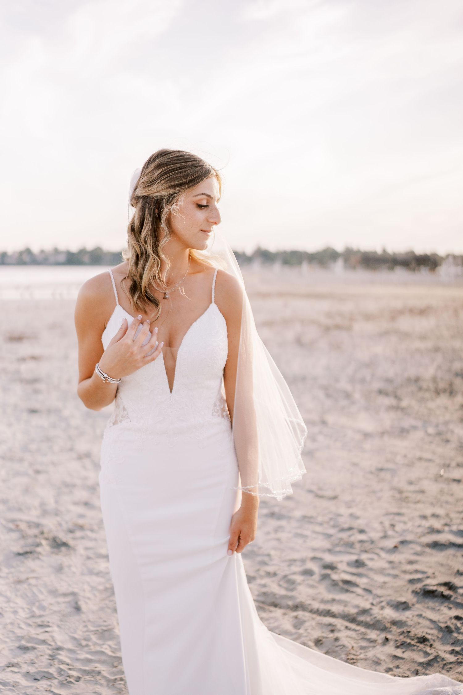 A bride in a flowing white dress poses elegantly on a sandy beach during sunset.