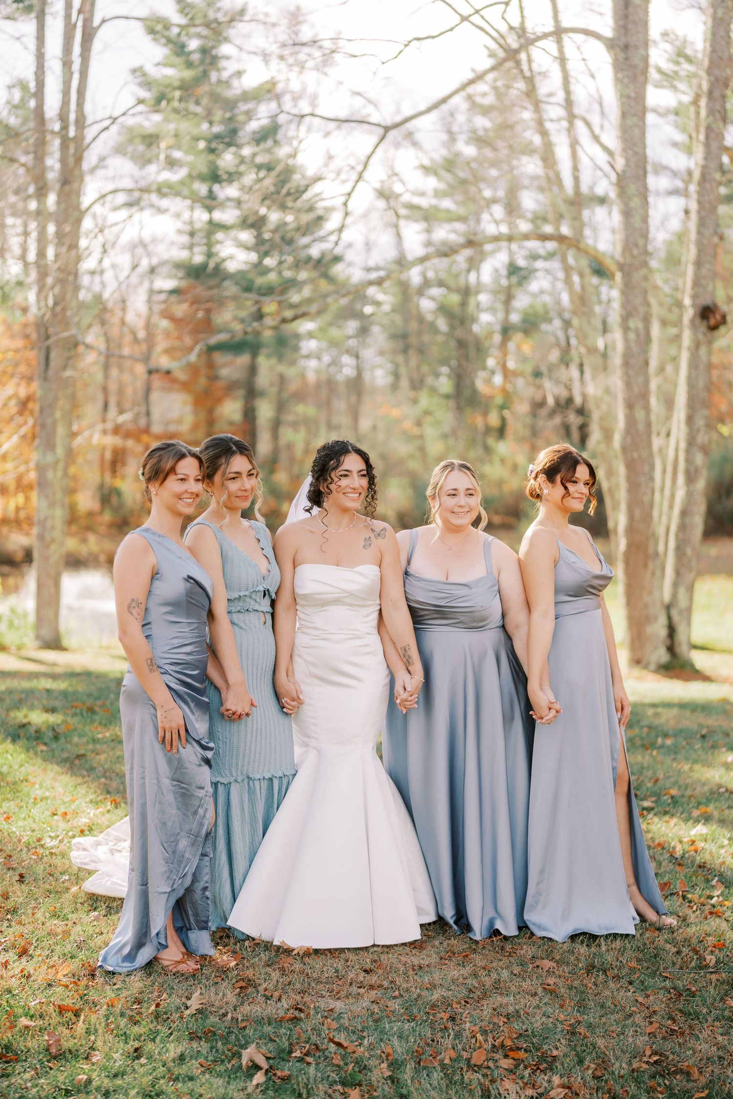 Wedding party in dusty blue bridesmaid dresses and white bridal gown pose together in autumn woods.