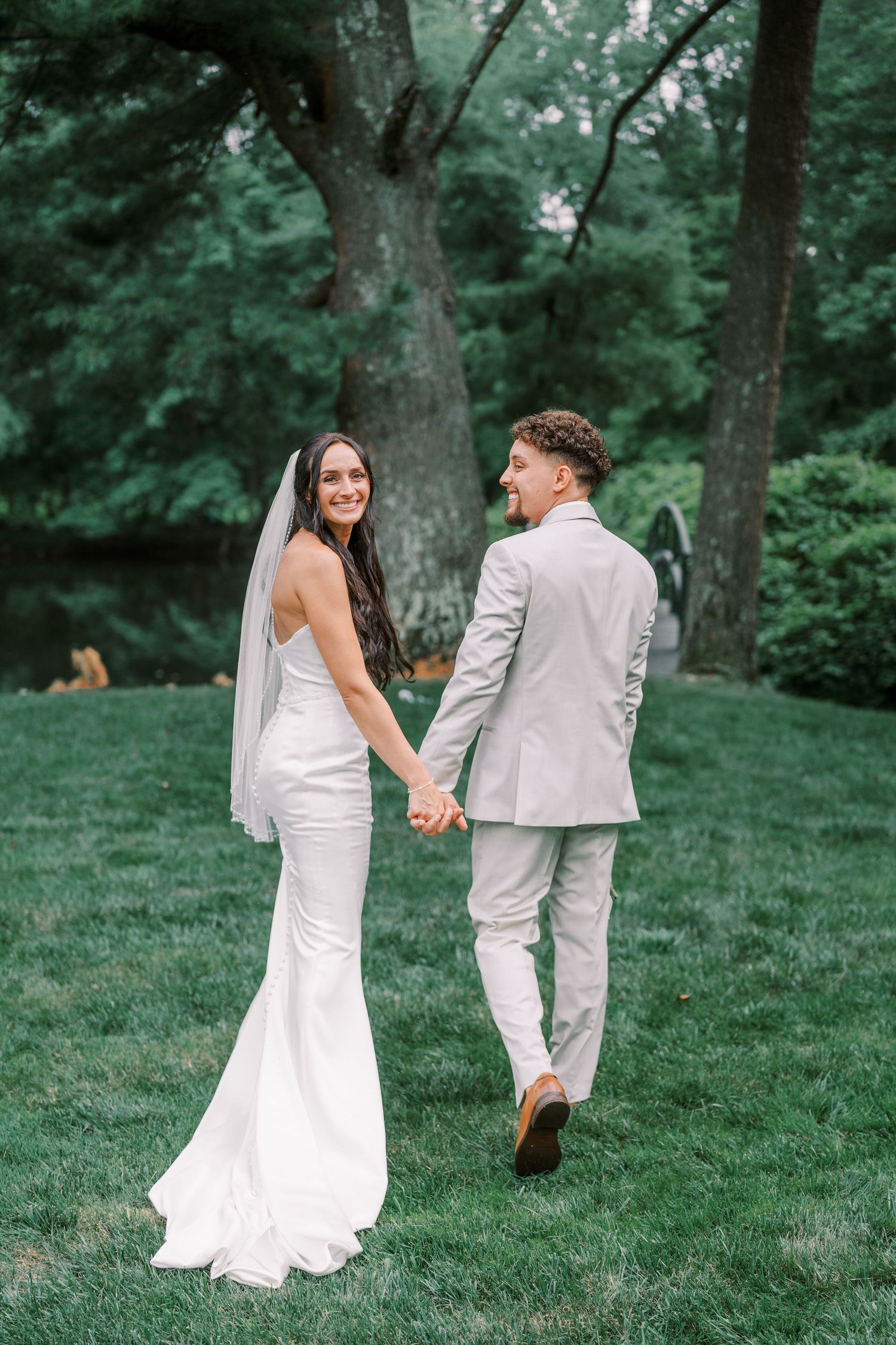 Wedding couple strolls together on grassy lawn with trees, bride's white dress trailing behind them.