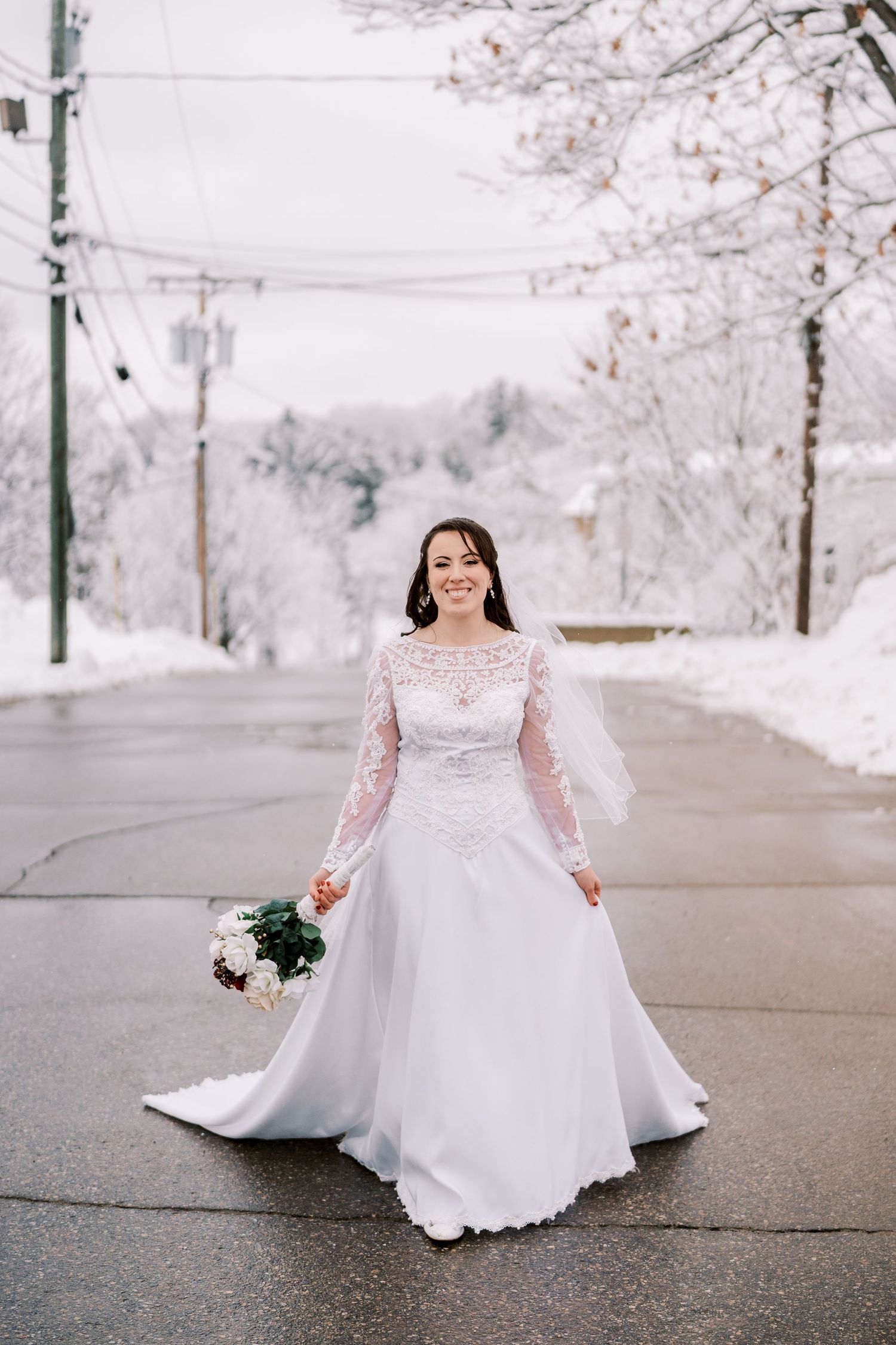 A bride in a long-sleeved lace wedding gown poses on a snow-covered street.