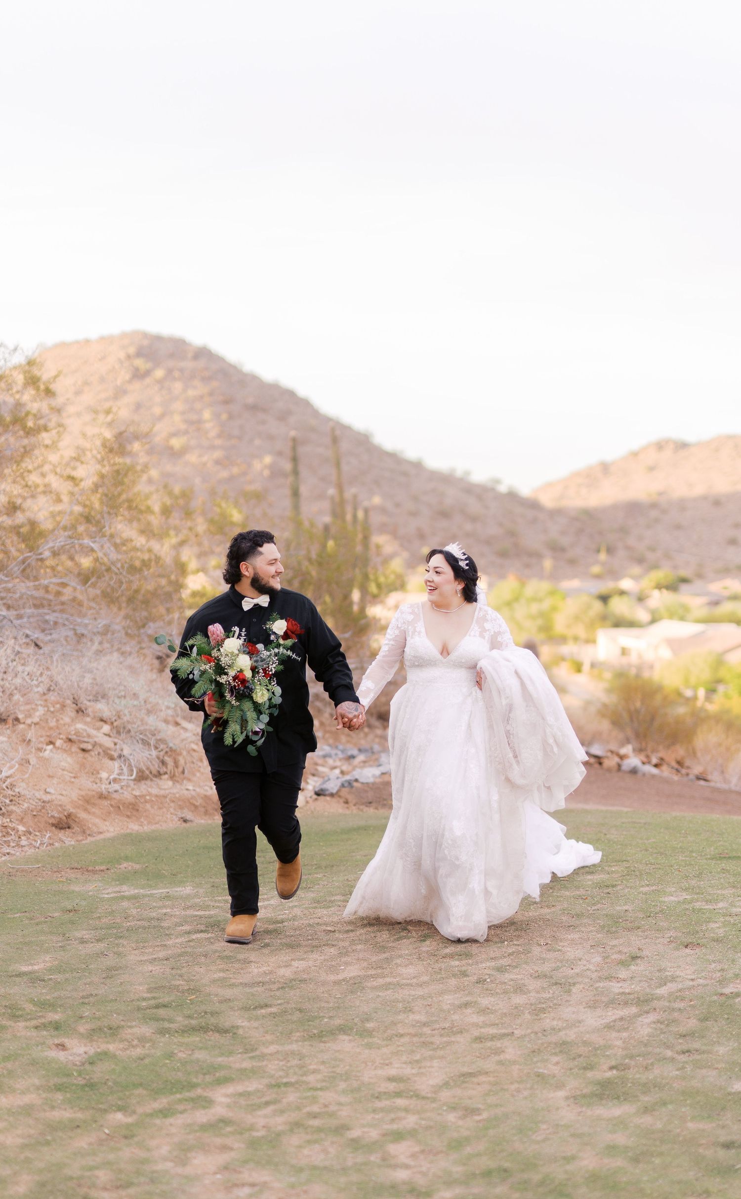 A couple runs hand in hand on grassy terrain with desert mountains in the background at sunset during their wedding celebration.
