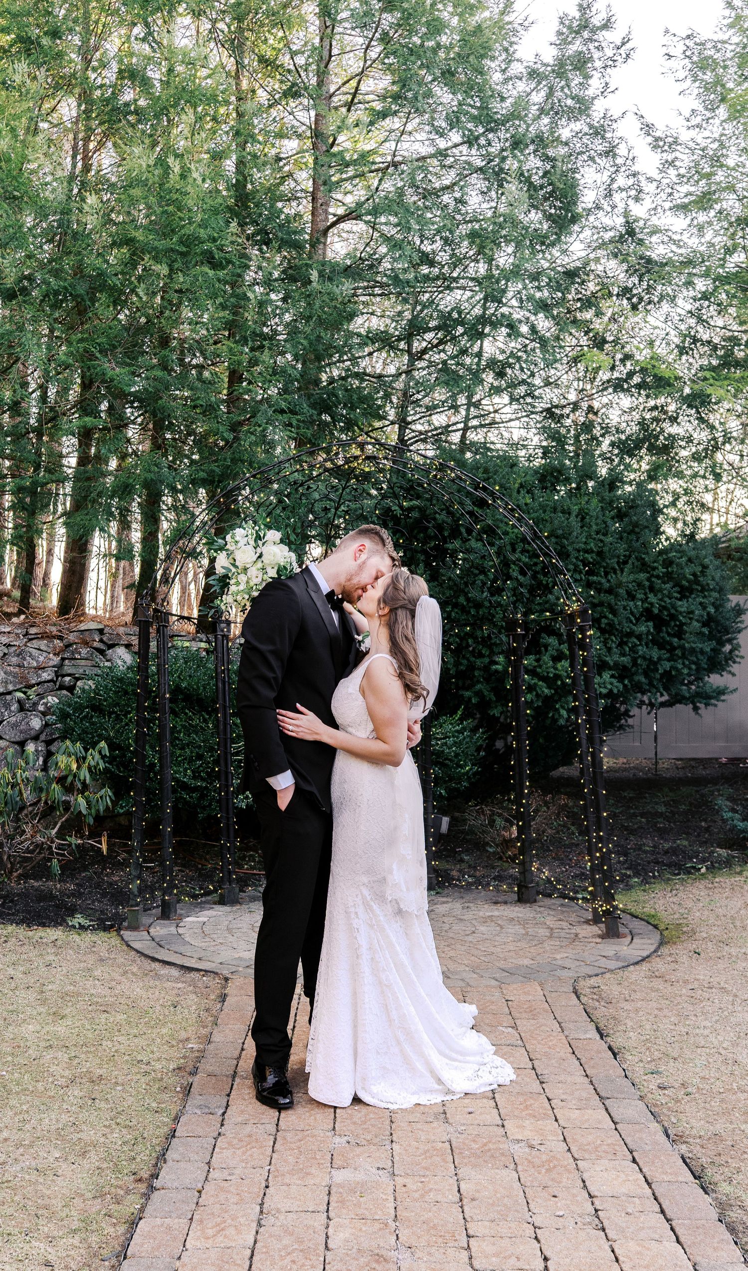 Romantic wedding couple shares intimate moments on garden brick pathway surrounded by evergreen trees.