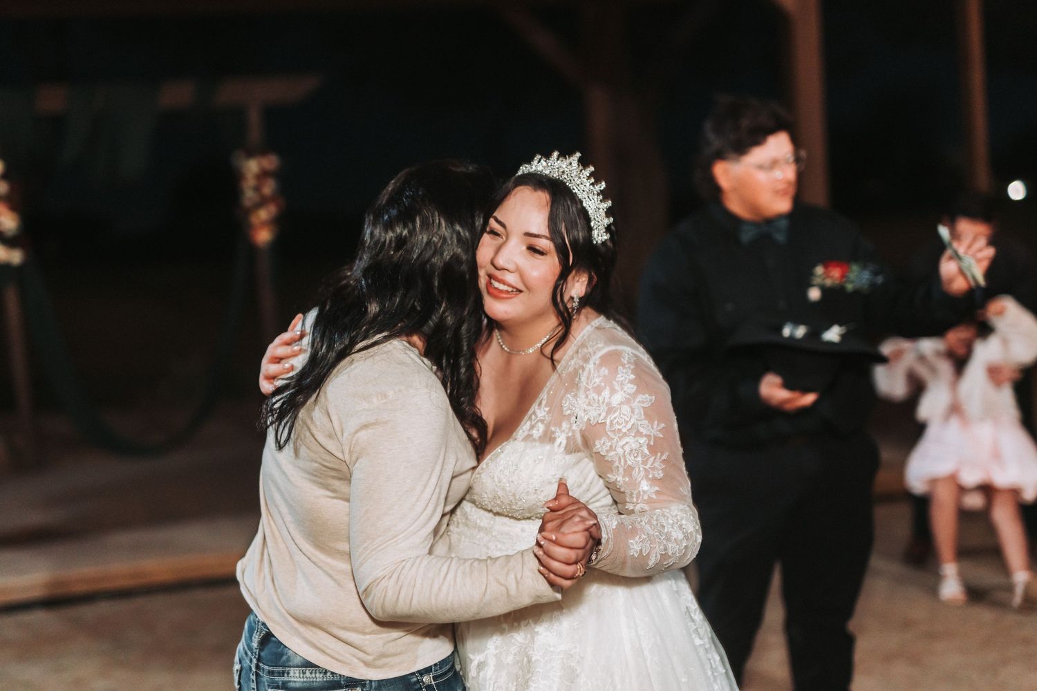 Two people embrace warmly on a dance floor during a wedding celebration while others mingle in the background.