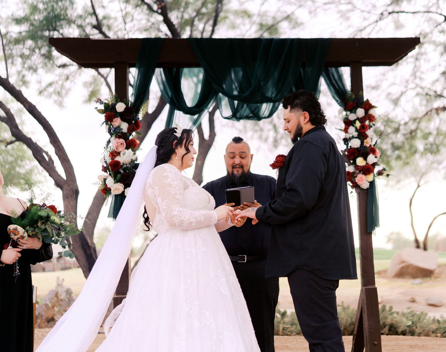 An outdoor wedding ceremony takes place beneath a wooden pergola decorated with floral arrangements and draped fabric.