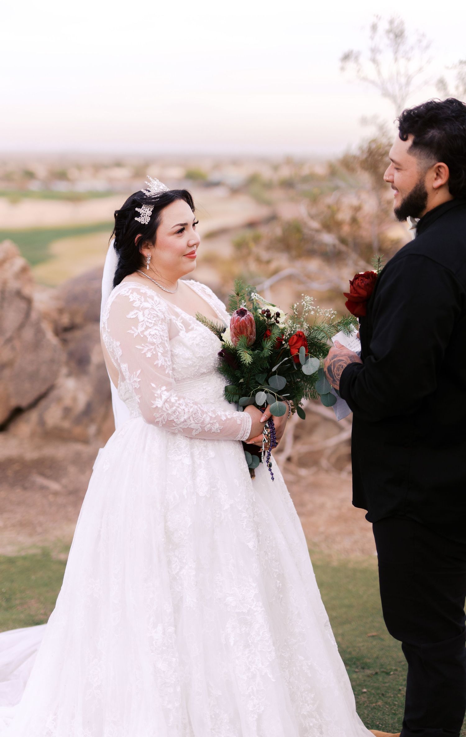 A couple shares a romantic moment on their wedding day at sunset, with the bride holding a red and white bouquet.