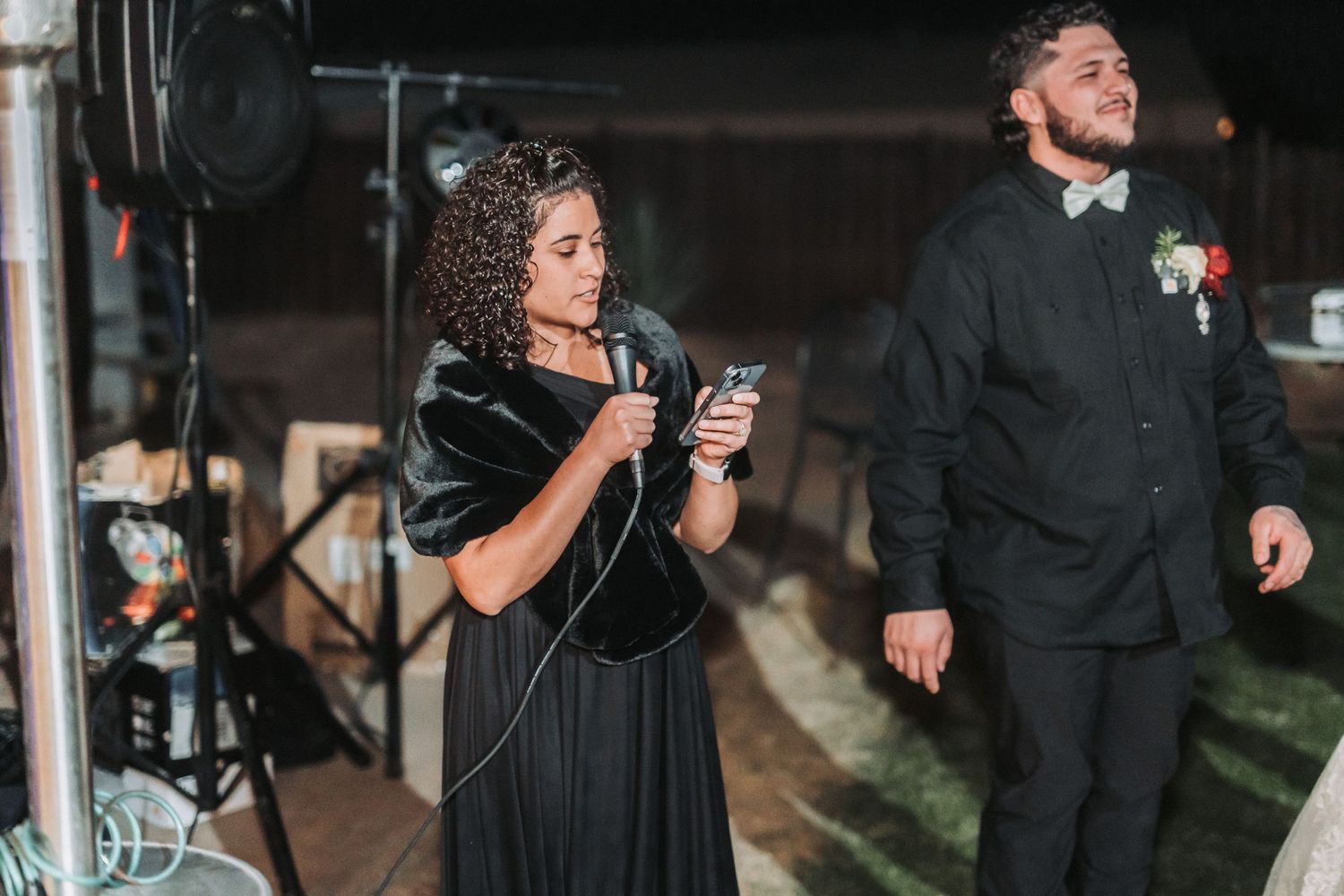 Two wedding guests at a nighttime reception with one speaking into a microphone in a dark dress while another stands nearby.