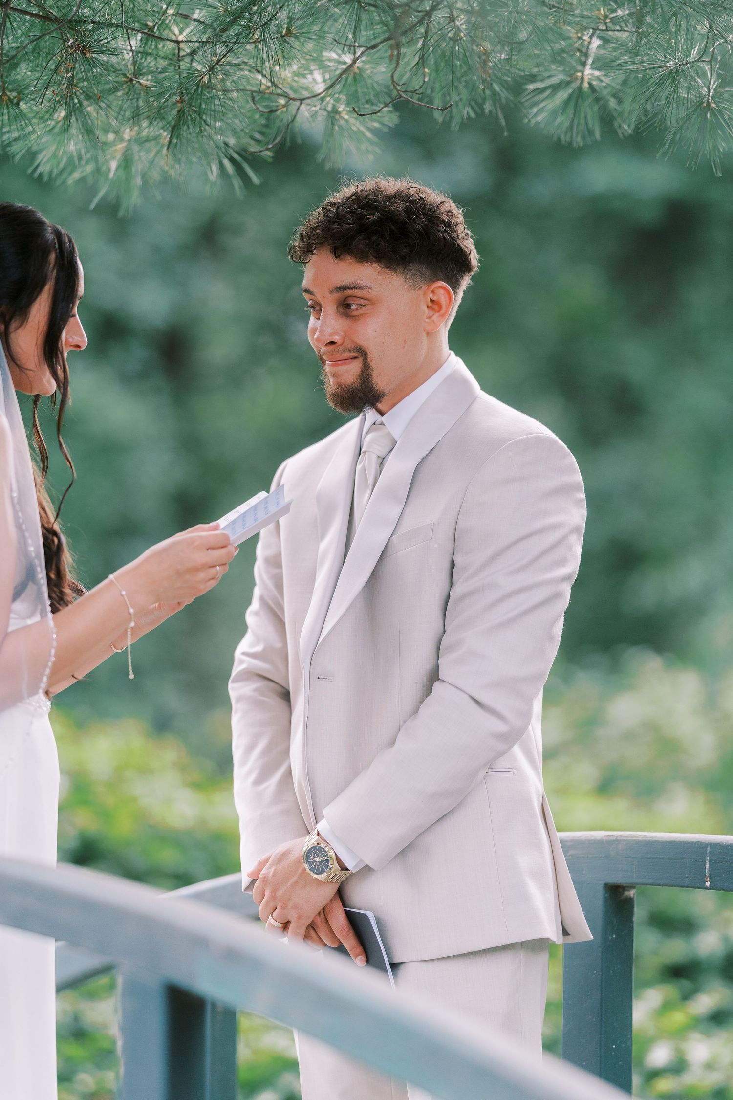 People in light colored formal attire standing on a deck or balcony with greenery in the background.