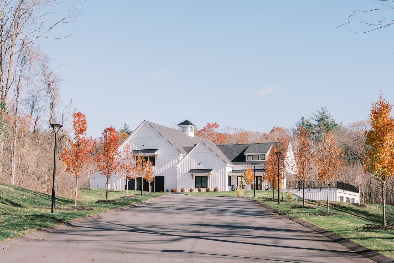 Modern farmhouse style venue with white exterior and black trim sits at end of paved driveway surrounded by autumn trees.