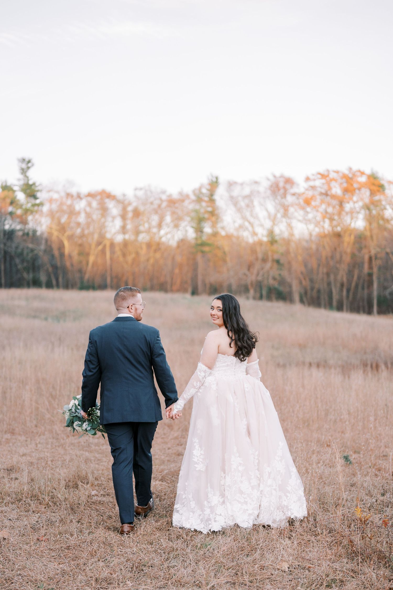 A couple in wedding attire walks through a golden autumn field at sunset holding hands.