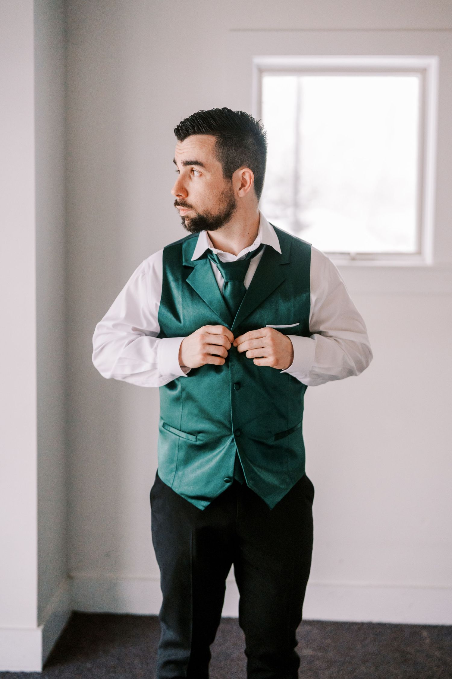 A person wearing a green vest and white dress shirt adjusts their attire while standing in a bright room.