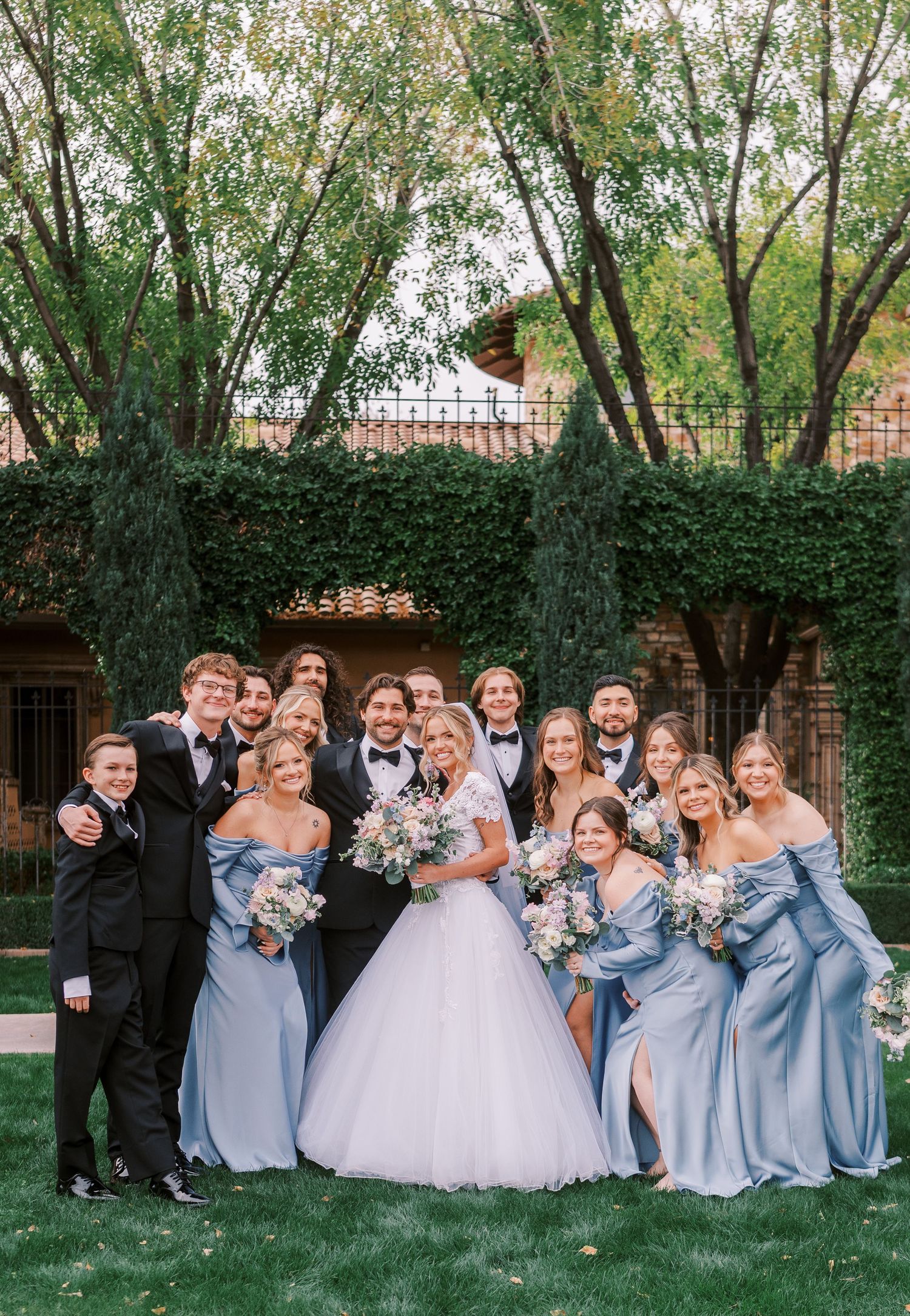 Wedding party in light blue bridesmaid dresses and formal attire pose together on grass near trees and brick building.